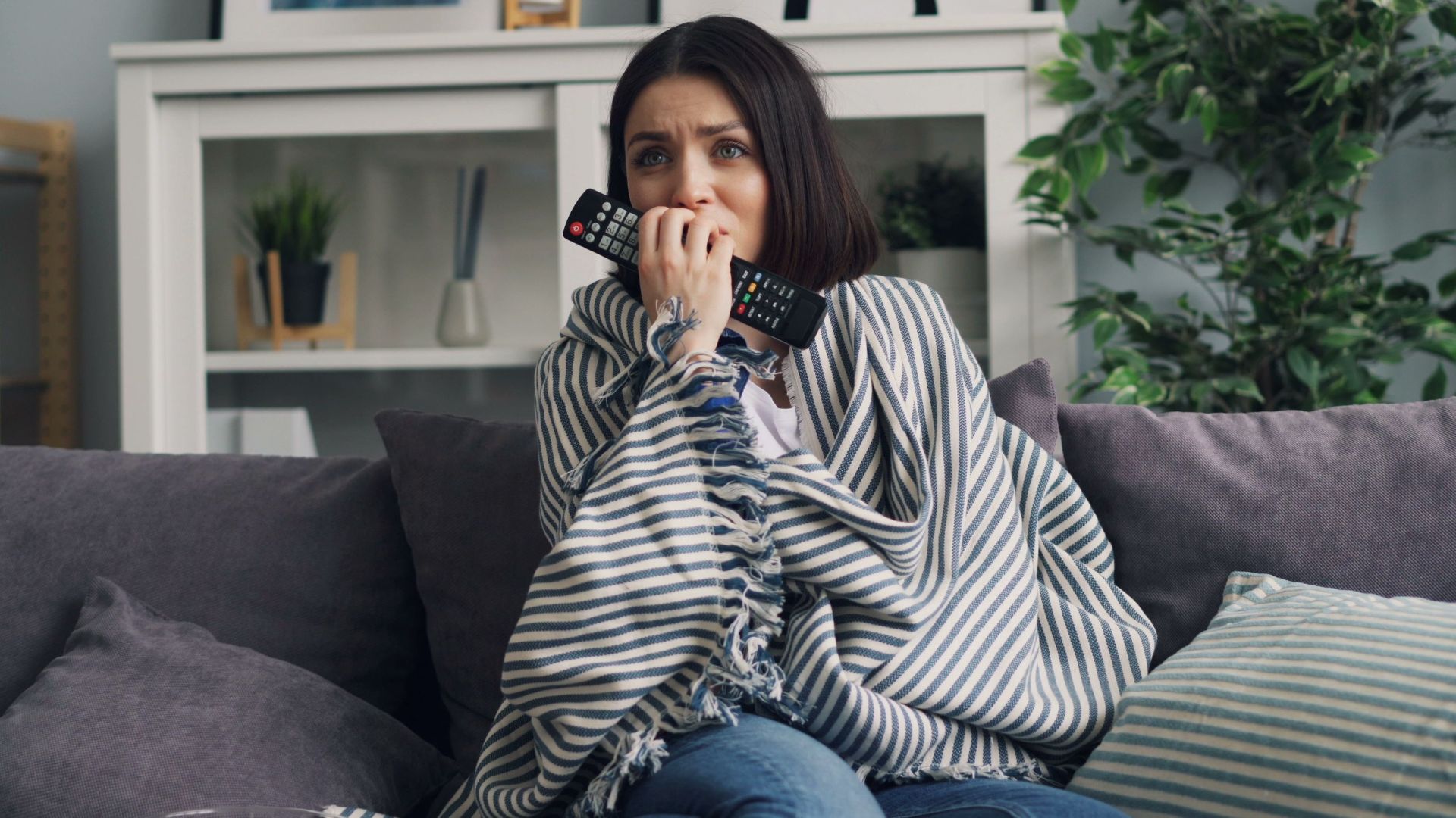 Anxious woman wrapped in a blanket watching TV at home with a remote in hand.