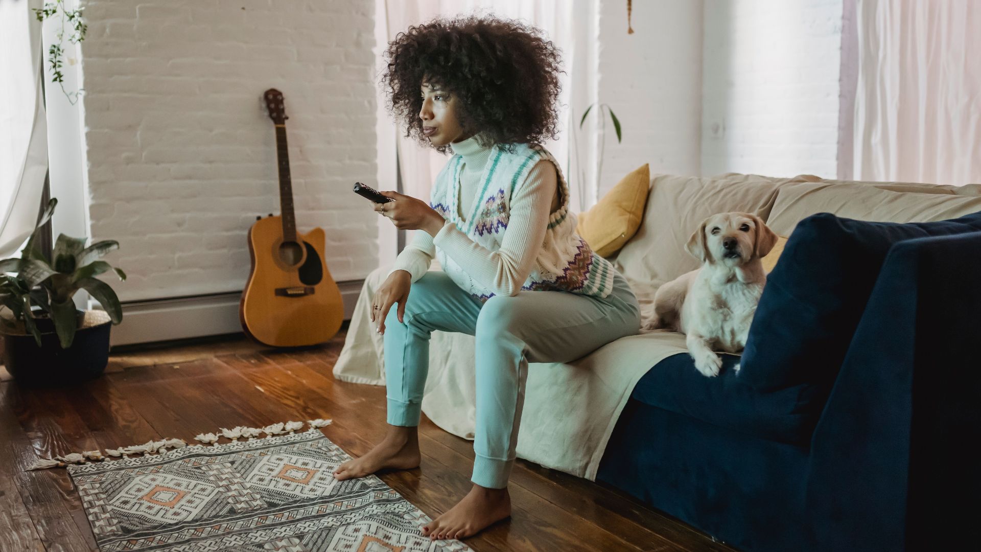 Full body of barefooted serious young African American lady with curly hair resting on sofa with obedient dog and switching TV channels with remote controller
