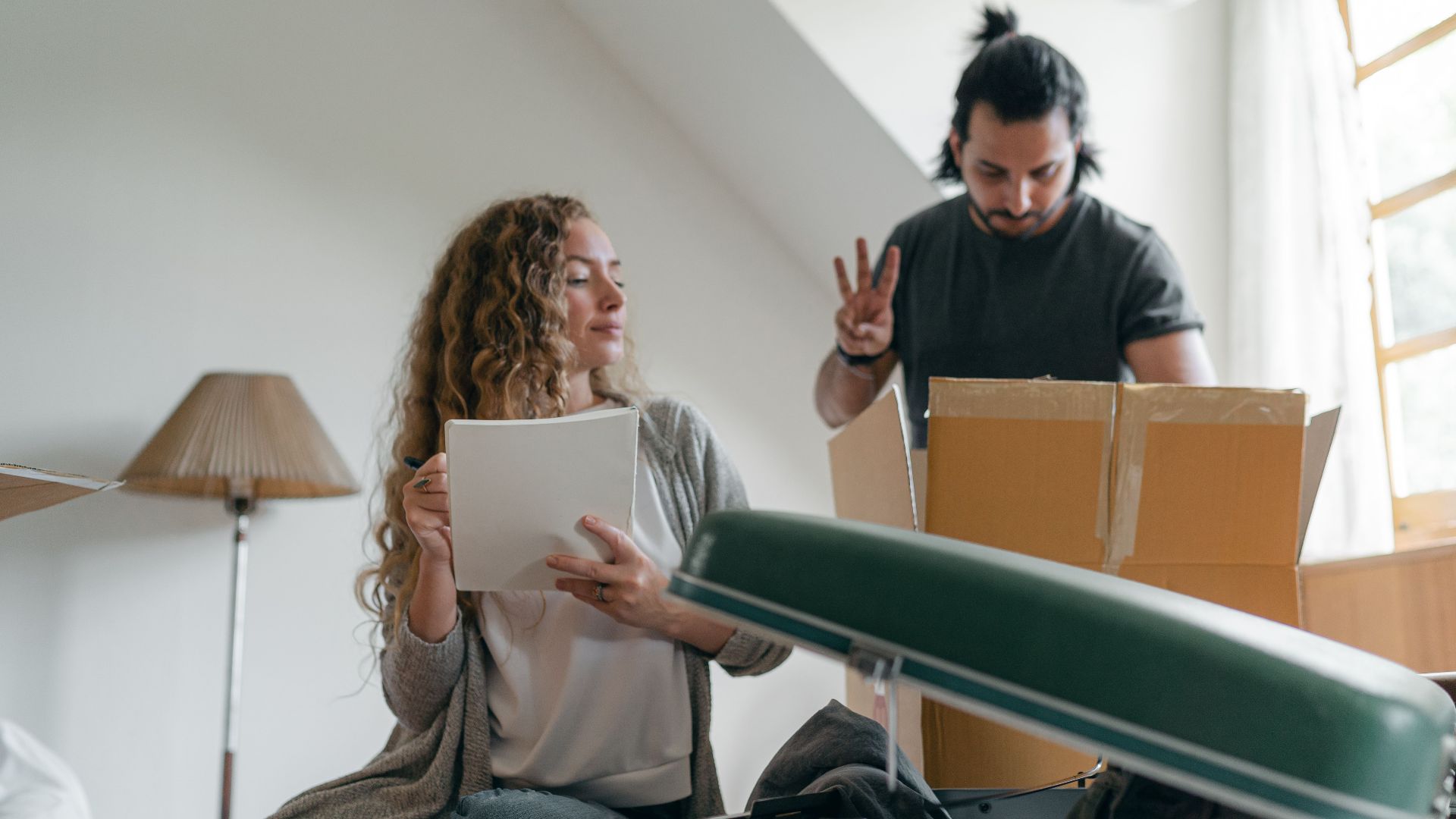 Positive concentrated couple counting belonging and taking notes while packing stuff into carton boxes and suitcases near window in sunny bedroom