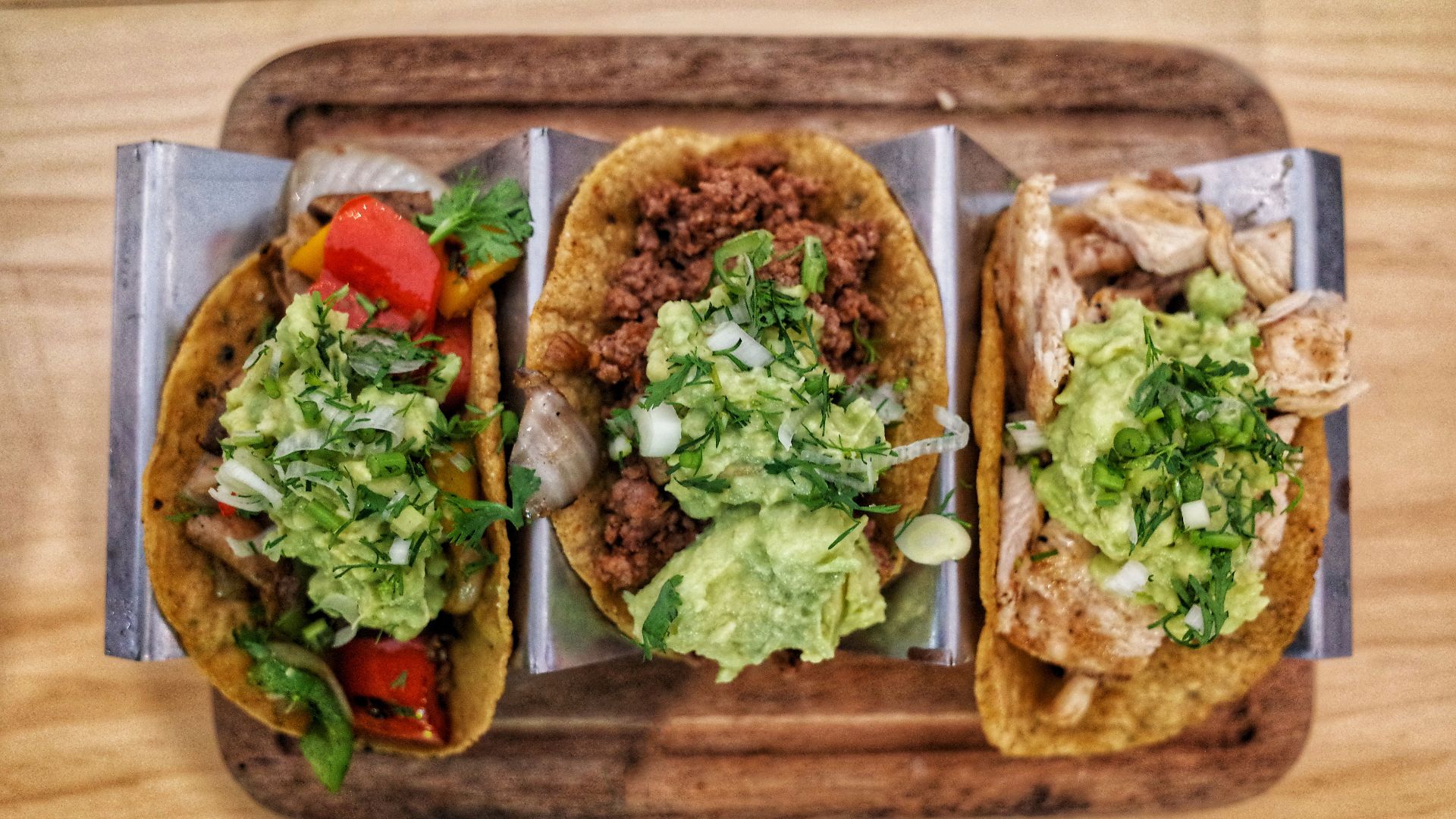 Three vibrant tacos with fresh guacamole, veggies, and meat on a wooden tray.