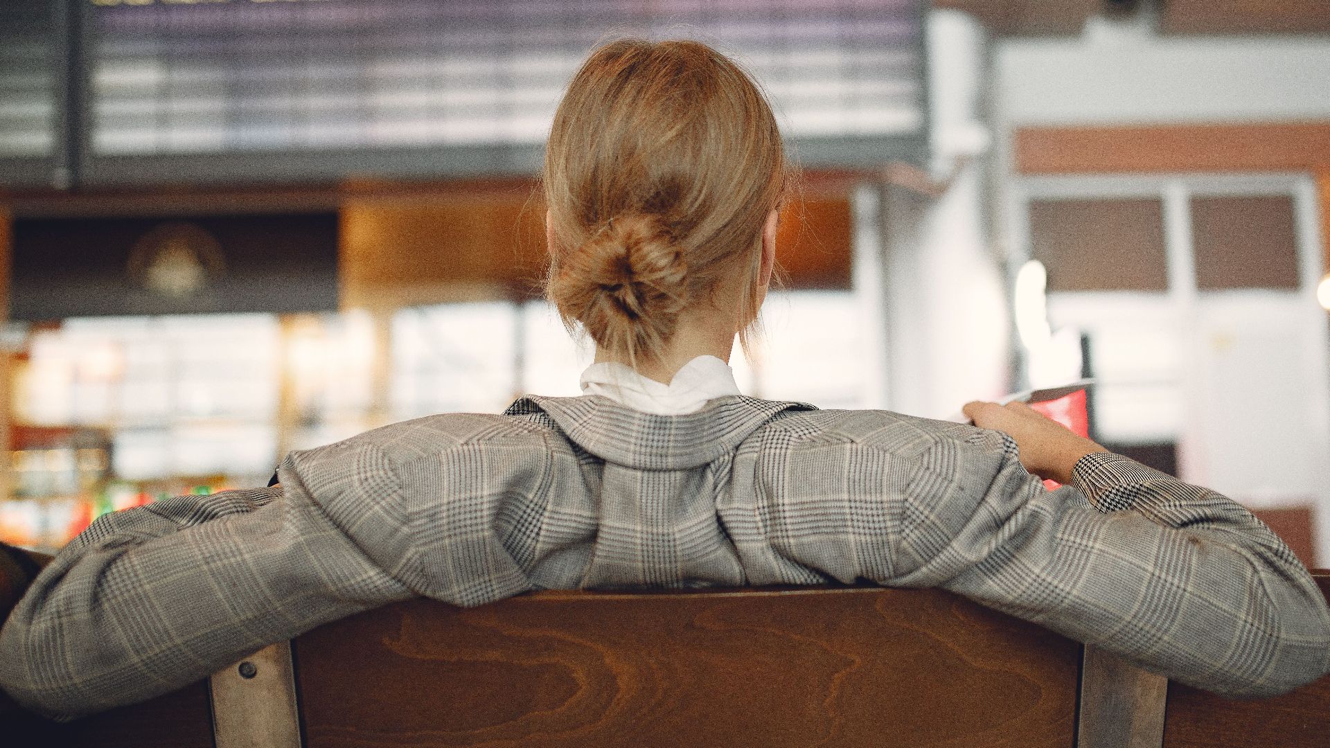 Back view of female employee in trendy jacket waiting for train departure and leaning on backs of wooden seats in train station
