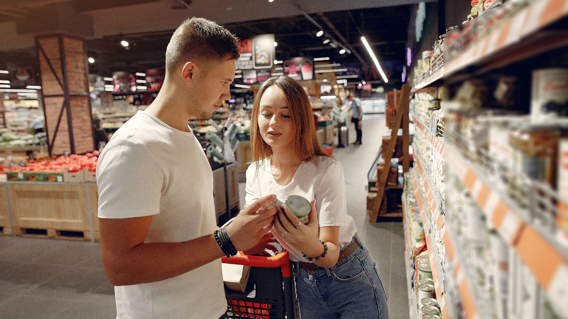 From above of young woman in casual wear showing canned food for man while standing with grocery trolley near shelves with products in modern supermarket