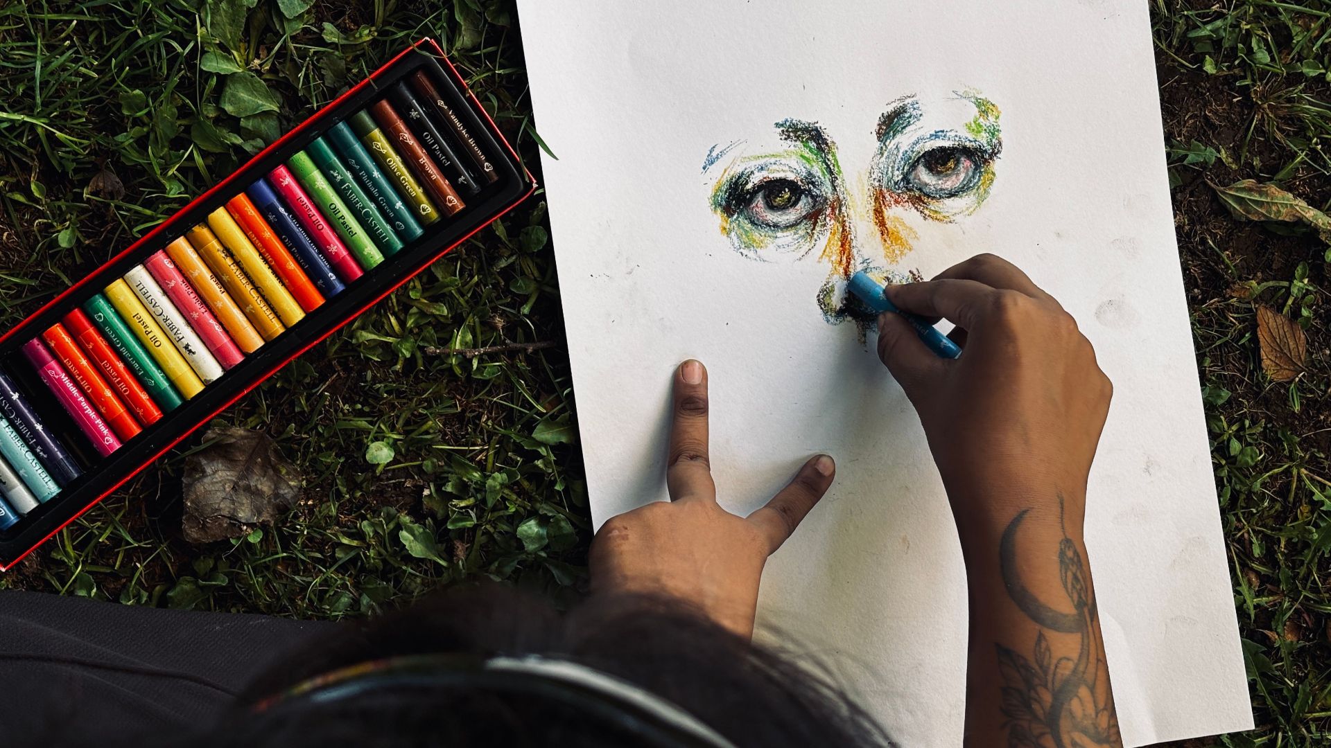 An artist drawing eyes on paper with pastels on a grass surface, showcasing creativity and color.