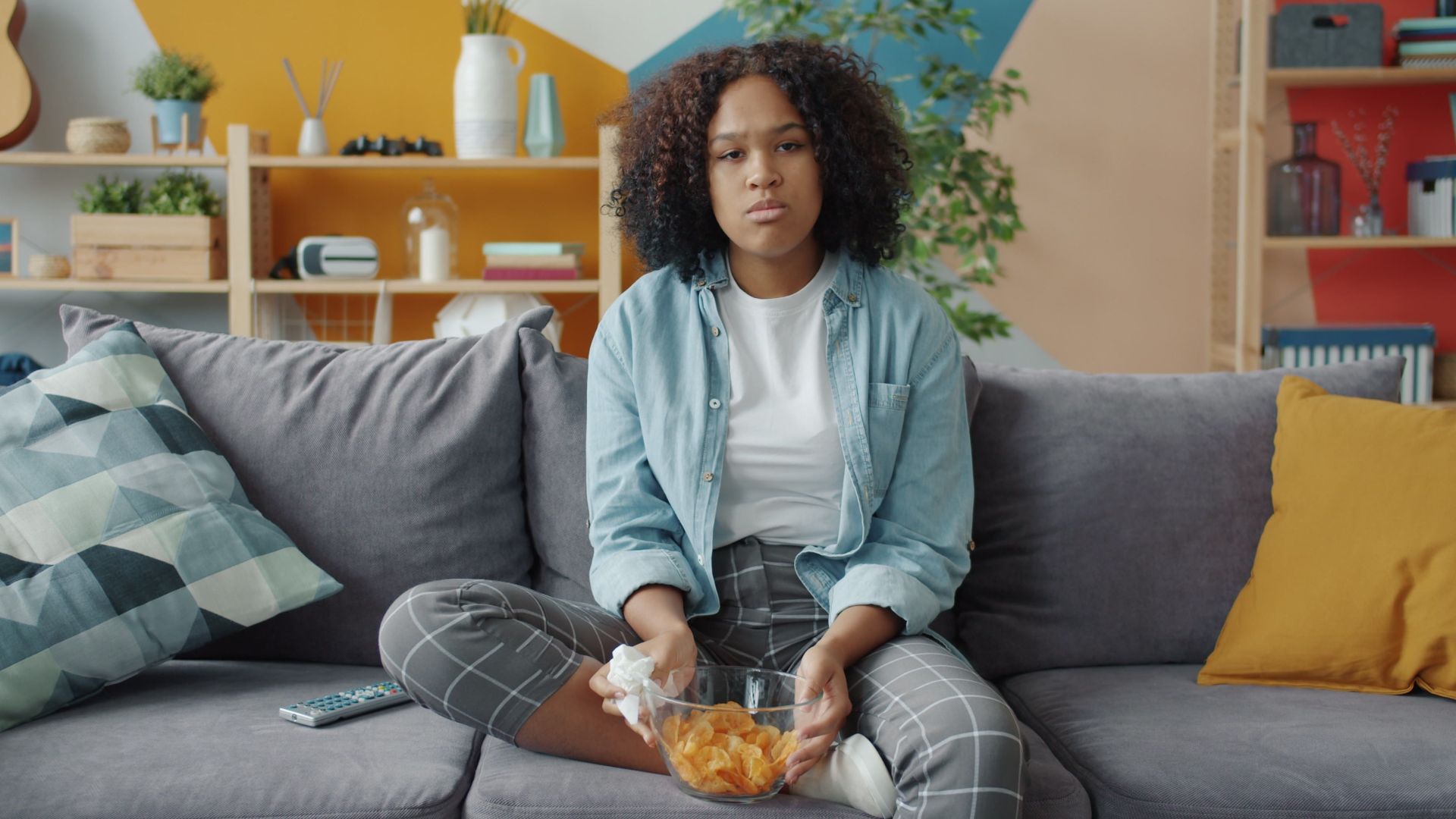 Young woman sitting on couch with snacks in a modern living room.