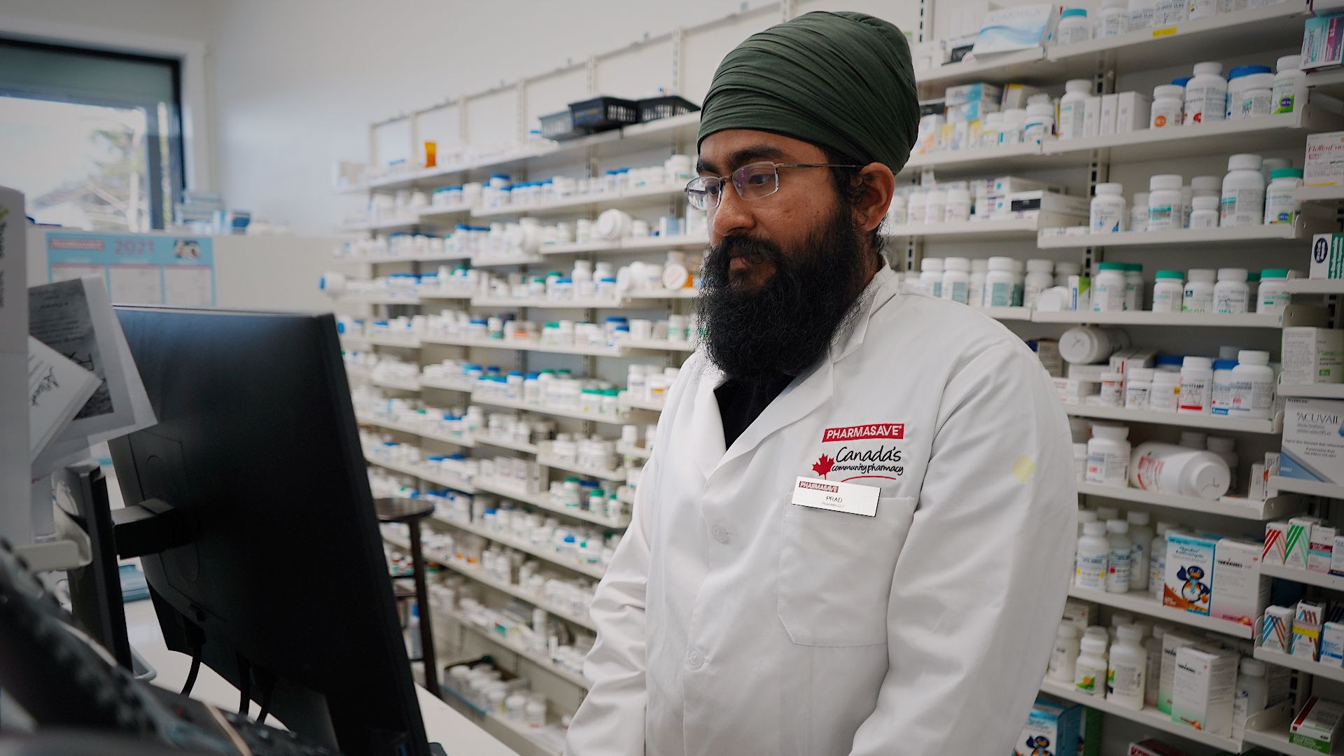 Pharmacist wearing a turban working at a computer in a pharmacy store with shelves filled with medicine.
