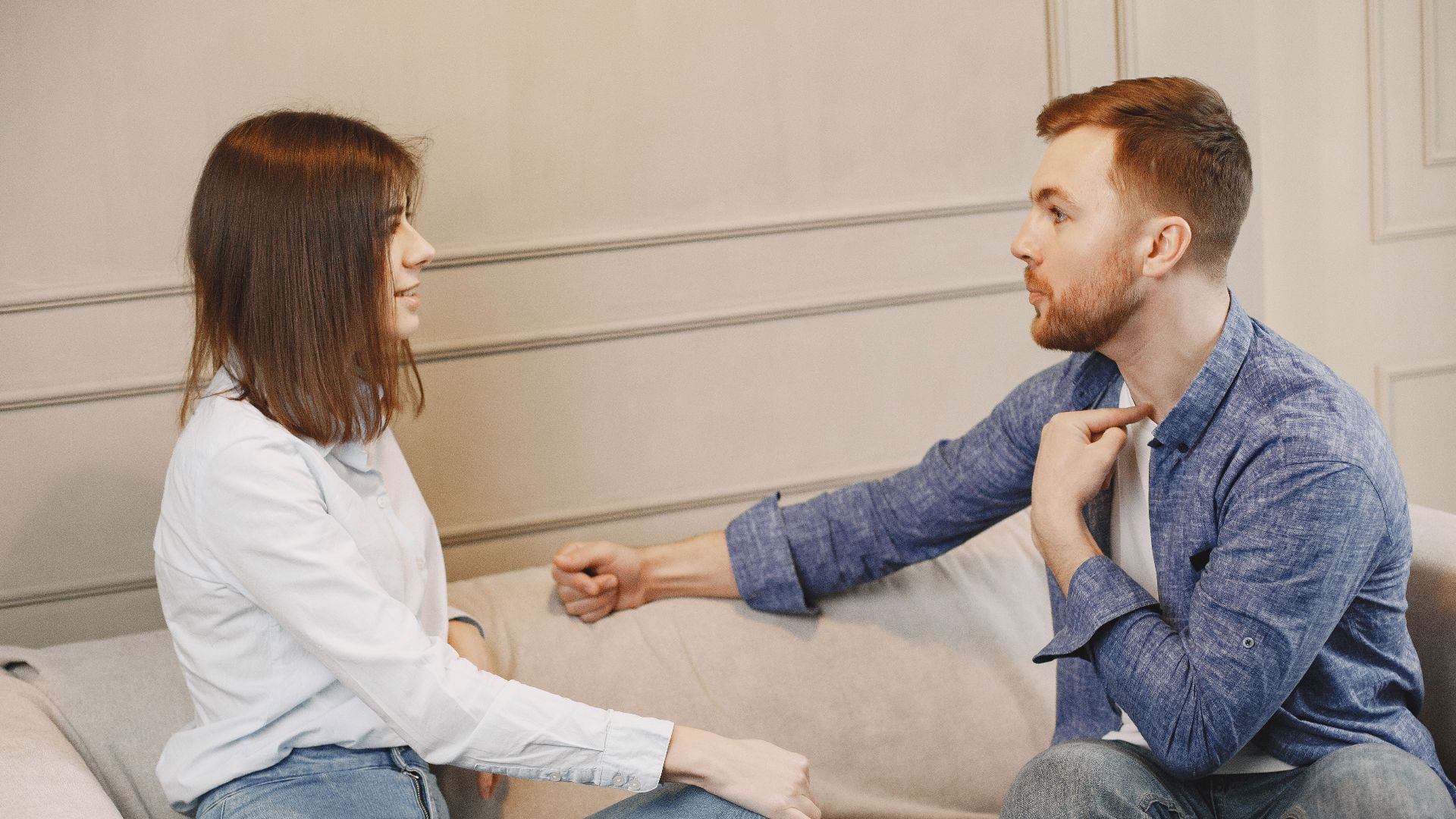 A young couple engaged in a conversation while sitting on a beige couch indoors.