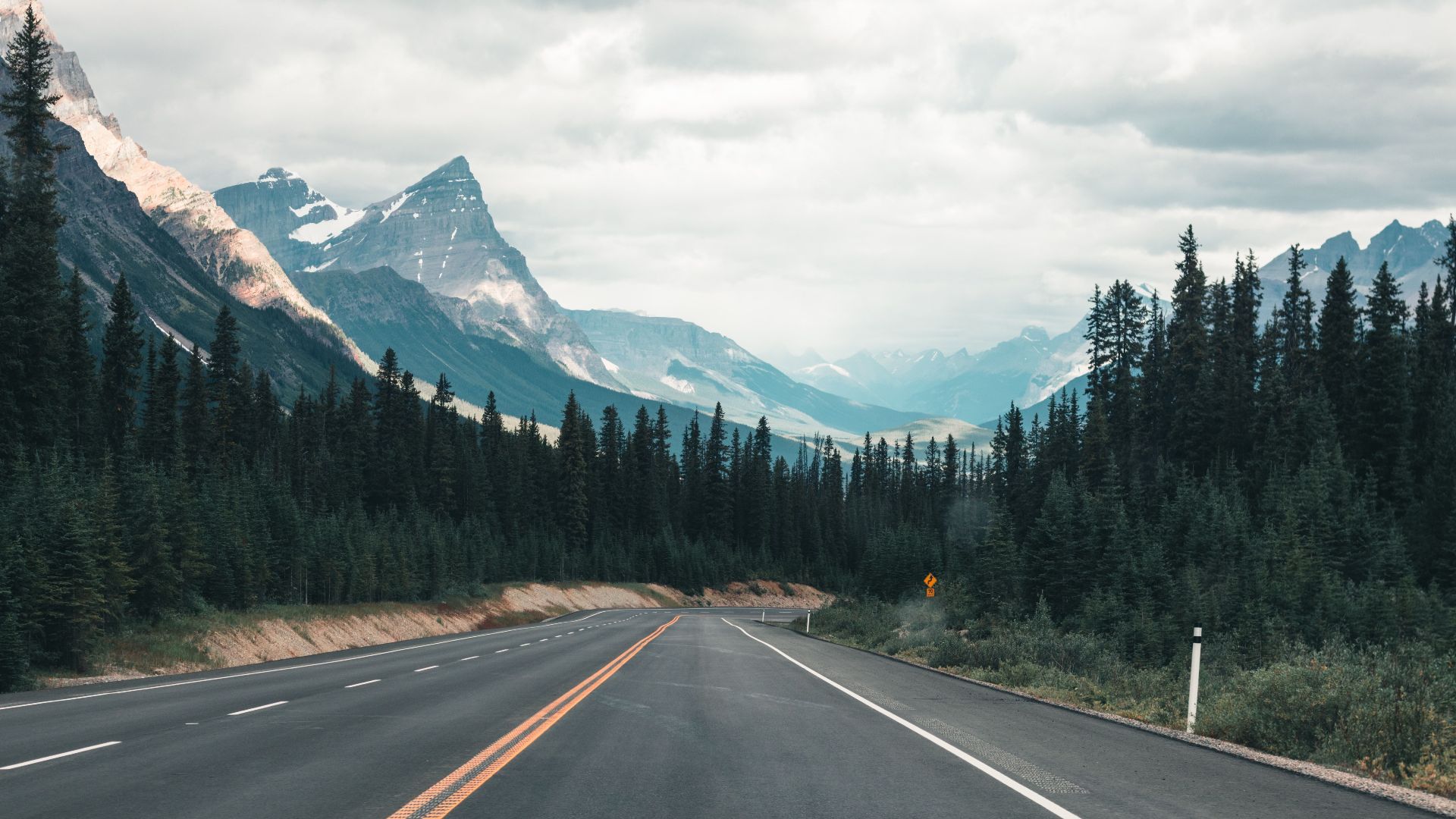 Empty highway through the majestic Canadian Rockies under a cloudy sky, framed by dense forest.