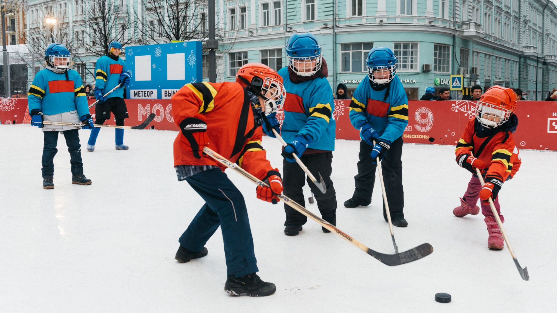 Kids in colorful gear playing ice hockey outdoors on a winter day.