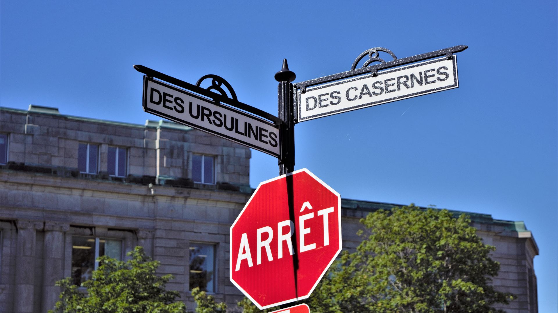 Bilingual street signs with stop sign against a clear blue sky in a city setting, featuring historical street names.