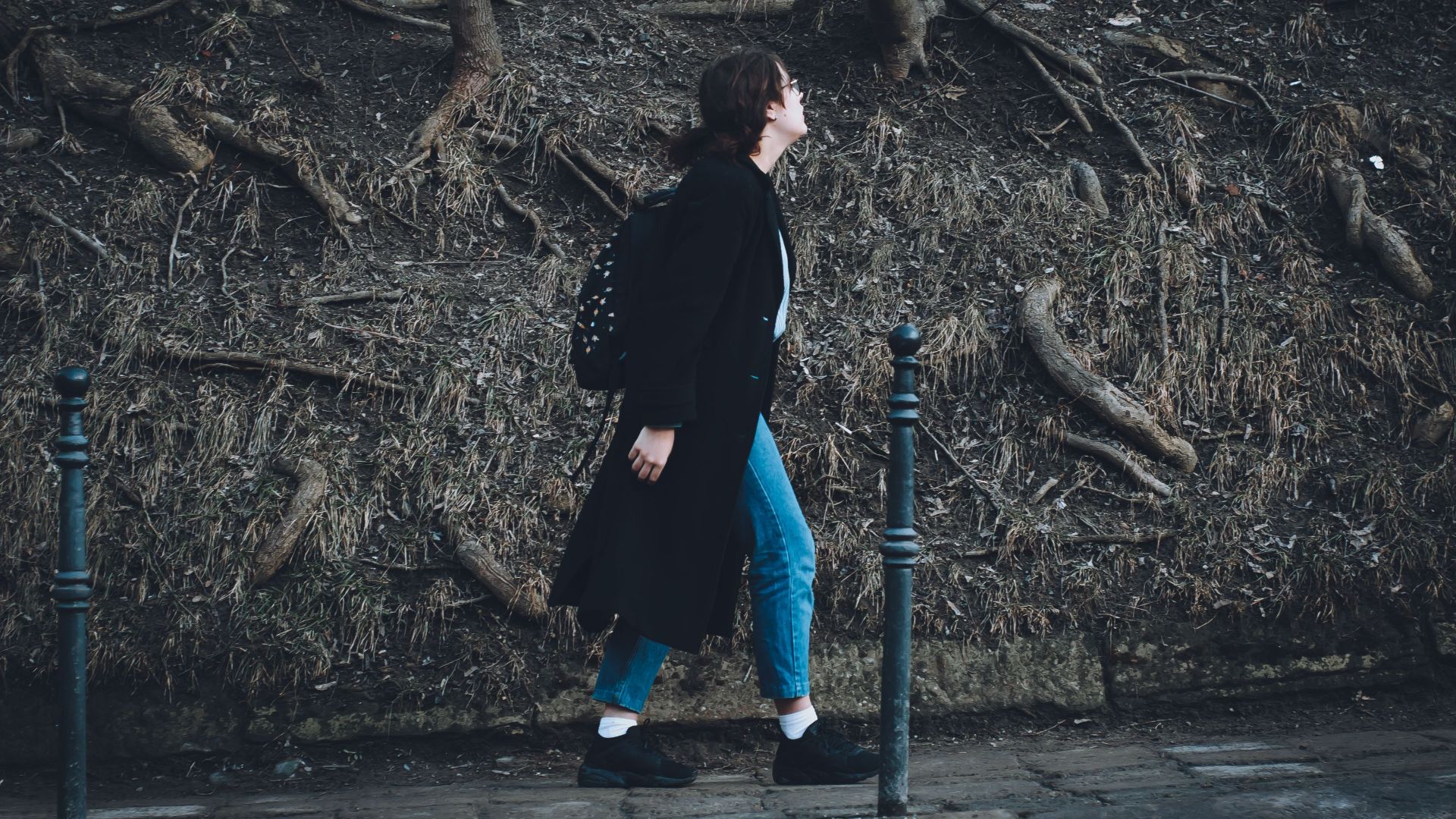 A woman in a long coat walks on a sidewalk surrounded by exposed tree roots.