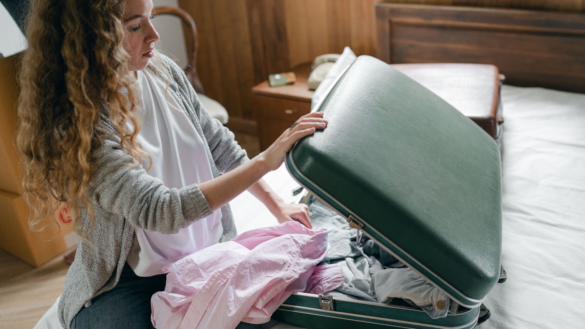 From above concentrated young female in casual wear with long curly hair sitting on bed and packing suitcase before trip