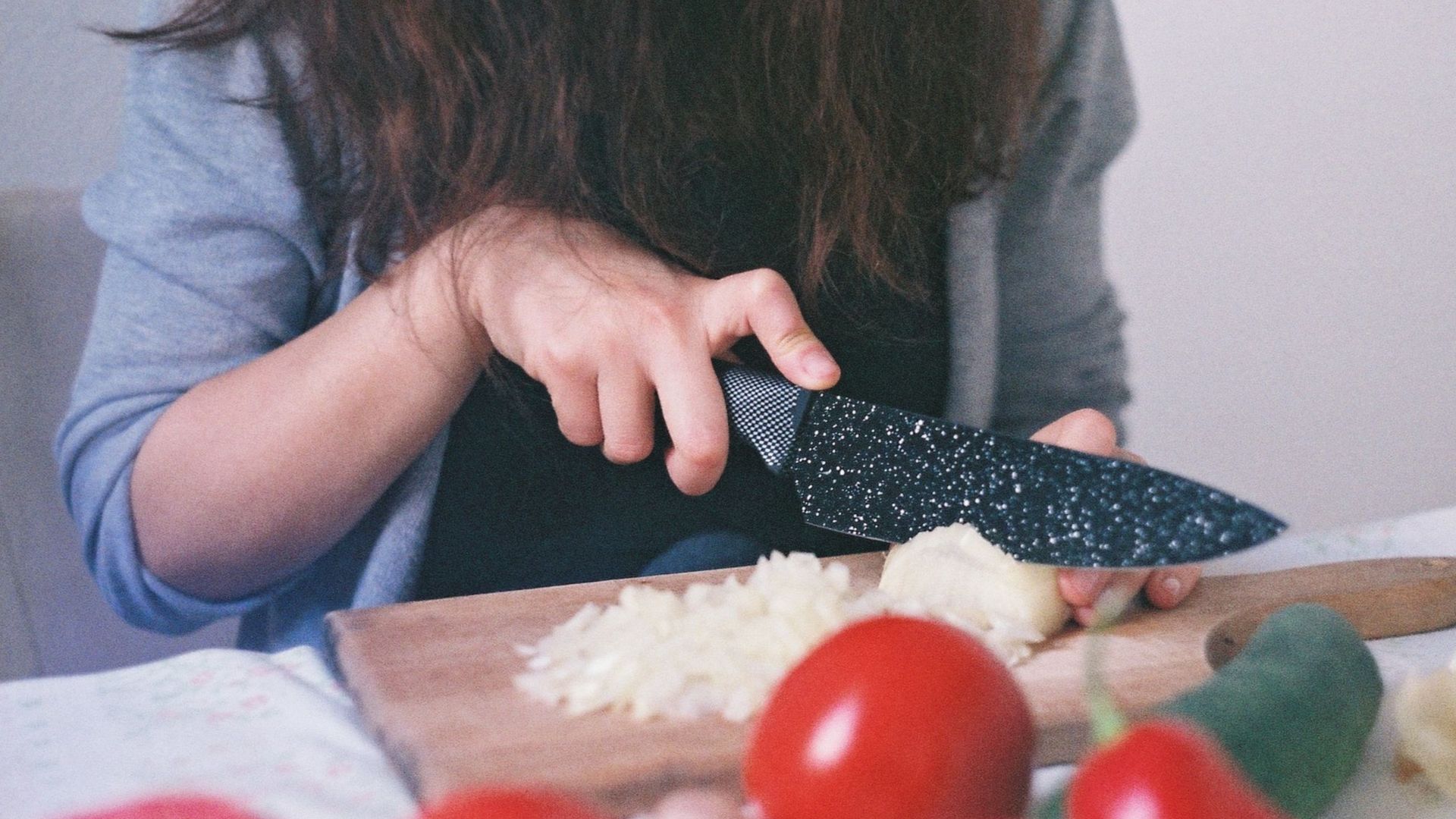 Close-up of a woman dicing onions and tomatoes on a wooden cutting board in a kitchen setting.