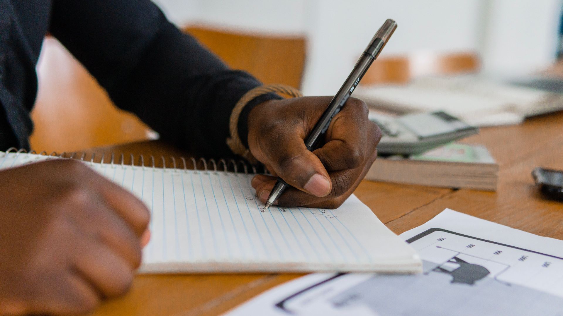 Close-up of hand writing on notebook at a wooden table, featuring paper and ballpen.