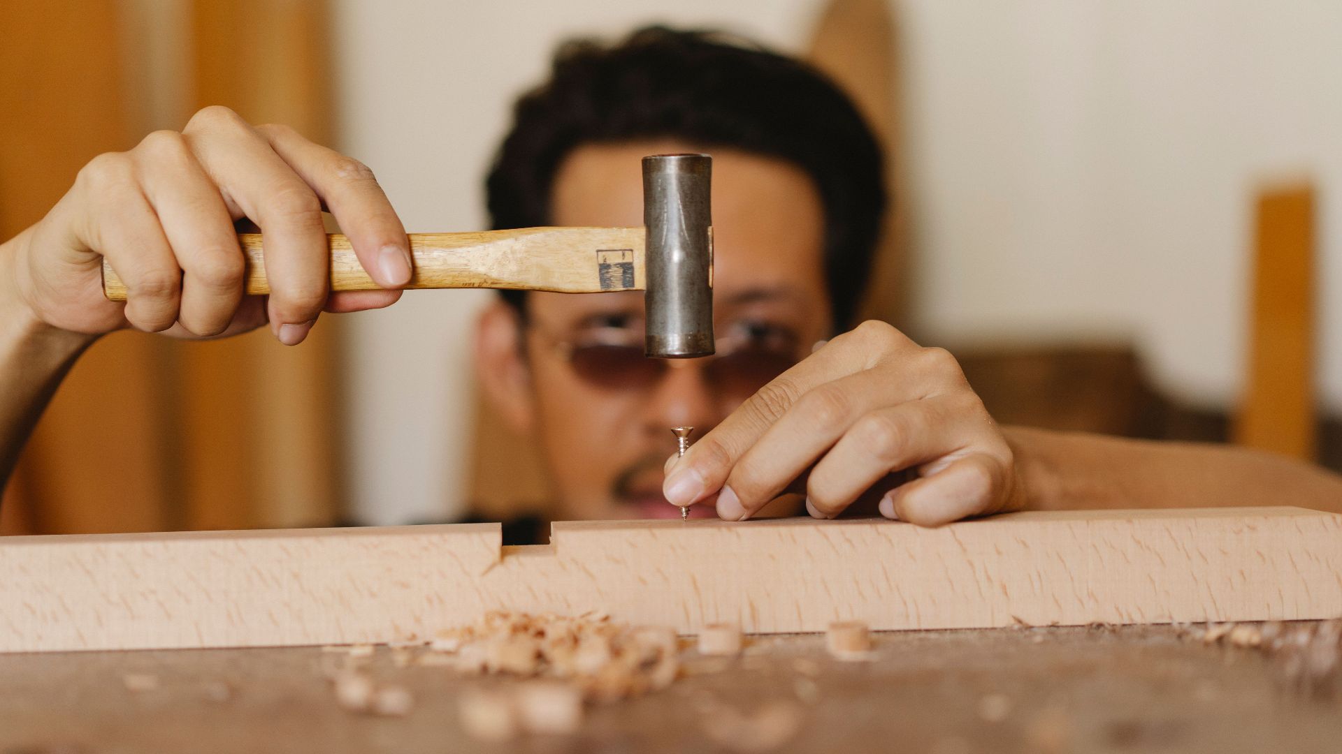 Concentrated young ethnic craftsman in eyeglasses hammering nail into wooden plank during work in carpentry studio