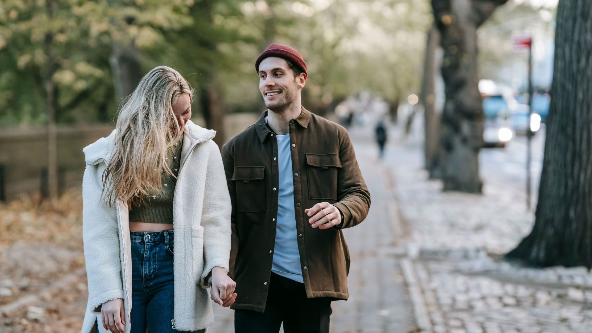 Positive young romantic couple in trendy outfits holding hands and smiling while walking on paved path on sunny autumn day