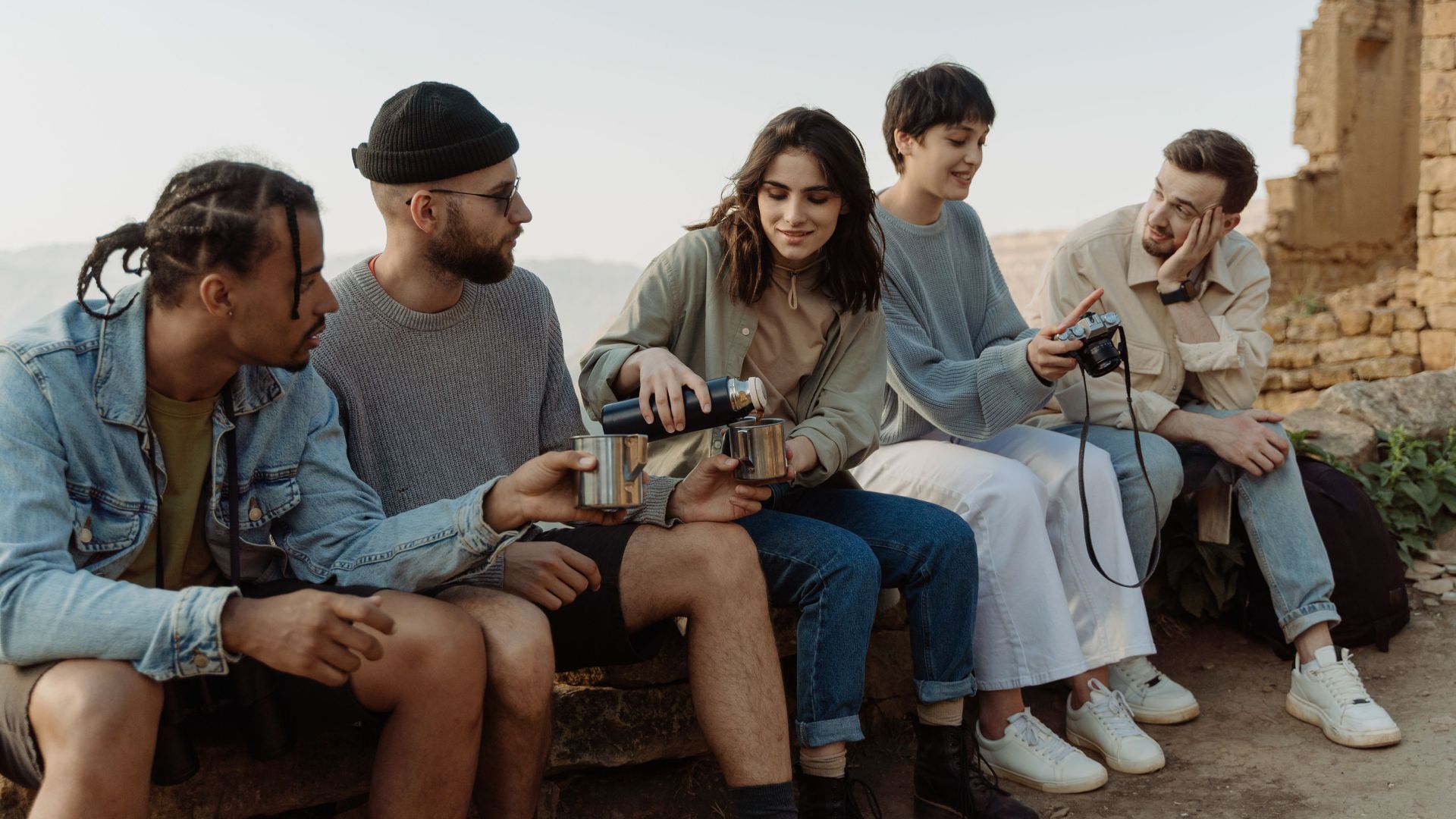 A diverse group of friends enjoy hiking together, sitting and sharing drinks outdoors on a sunny day.