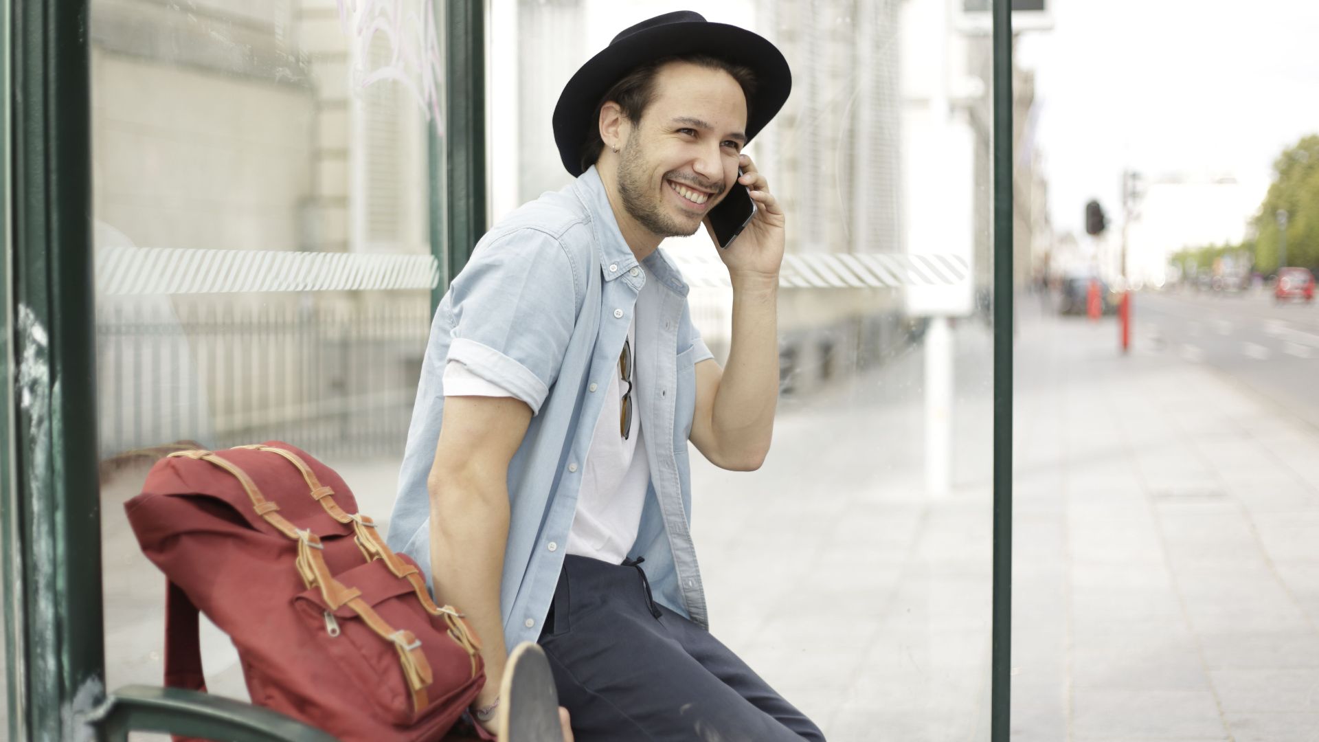 Casual young man sitting at a bus stop, using phone, and smiling in an urban environment.