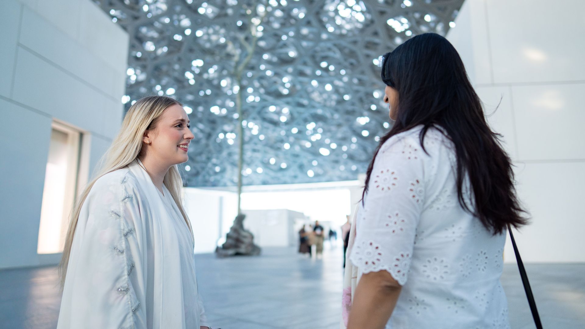 Two women enjoying conversation under the iconic Louvre Abu Dhabi architectural dome.