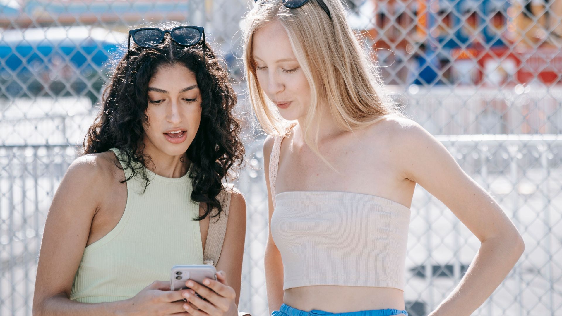 Two young women at an amusement park looking at a smartphone, standing by a fence in a sunny setting.