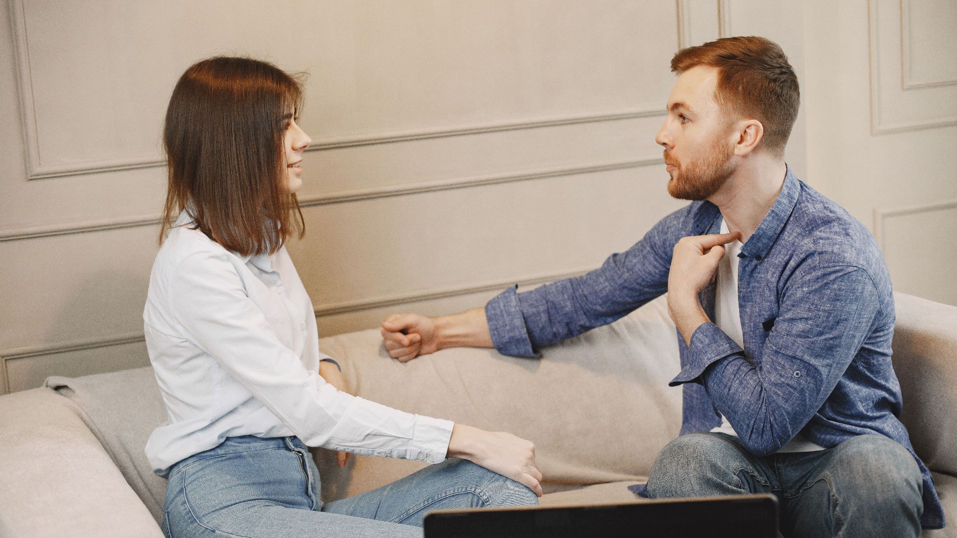 A young couple engaged in a conversation while sitting on a beige couch indoors.