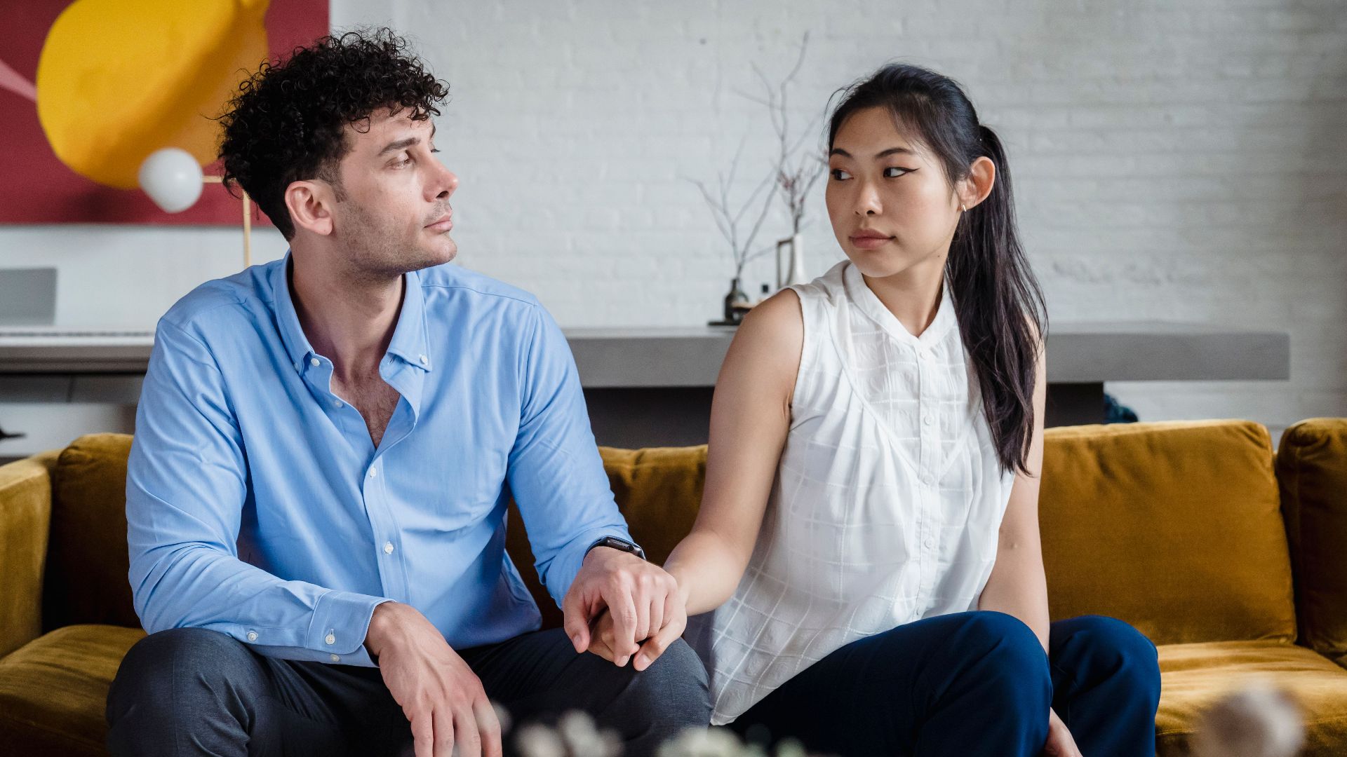 A couple sits on a sofa holding hands, showcasing intimacy and diversity indoors.