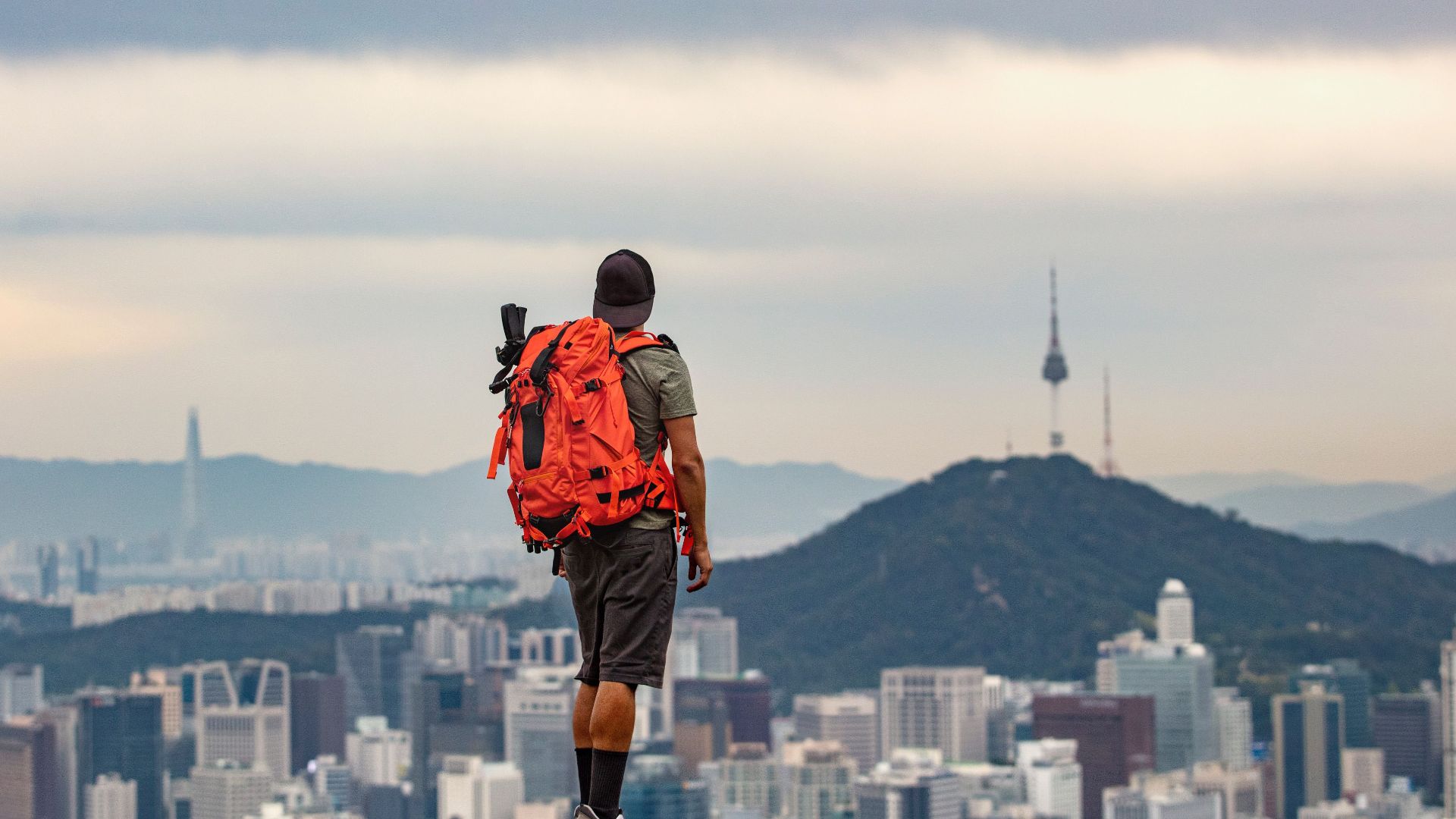Man with backpack observing Seoul cityscape from mountain peak.