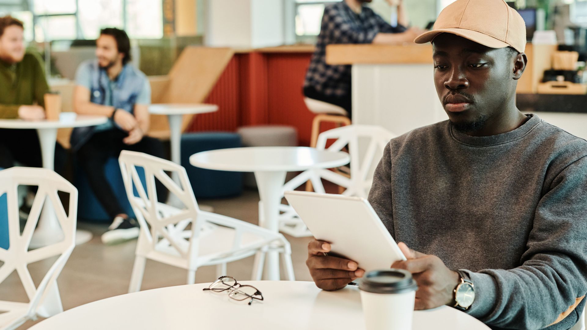 A focused young man in a cap uses a digital tablet in a contemporary café environment.