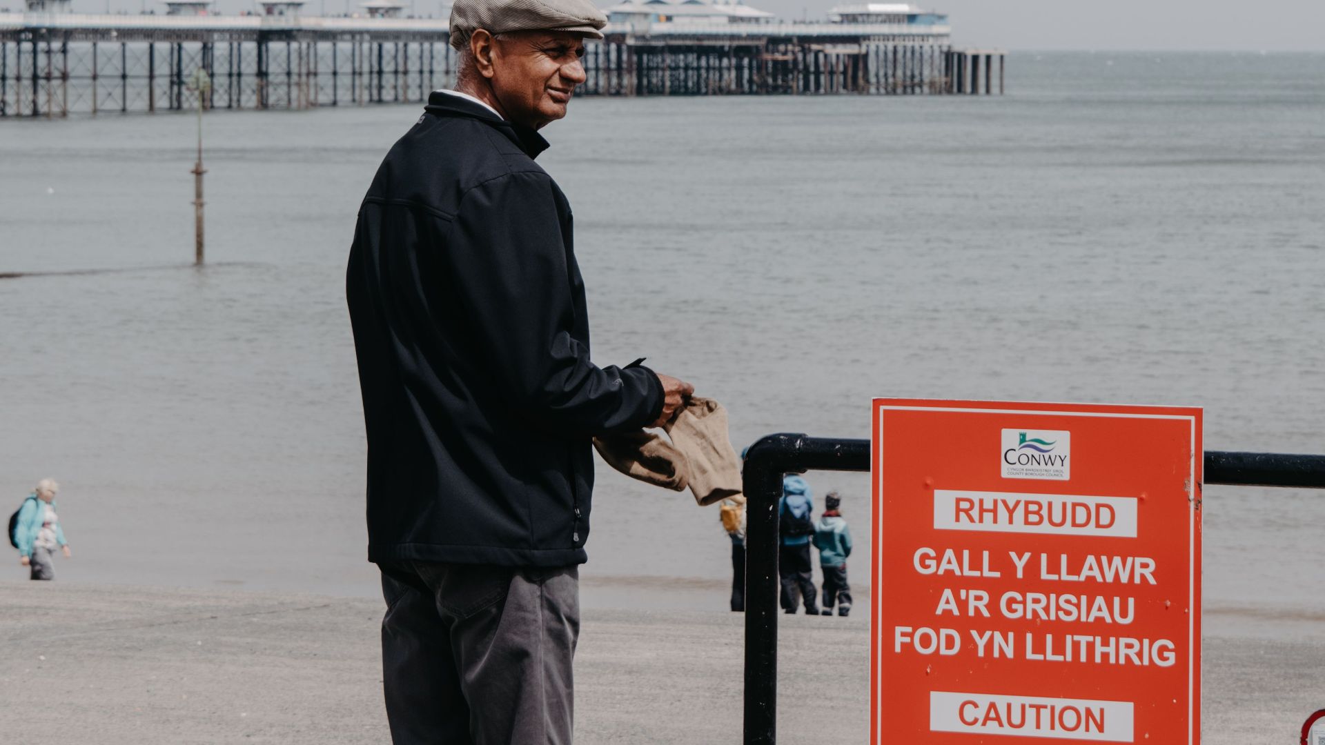 An elderly man stands near a caution sign by the seaside in Wales, with a pier in the background.