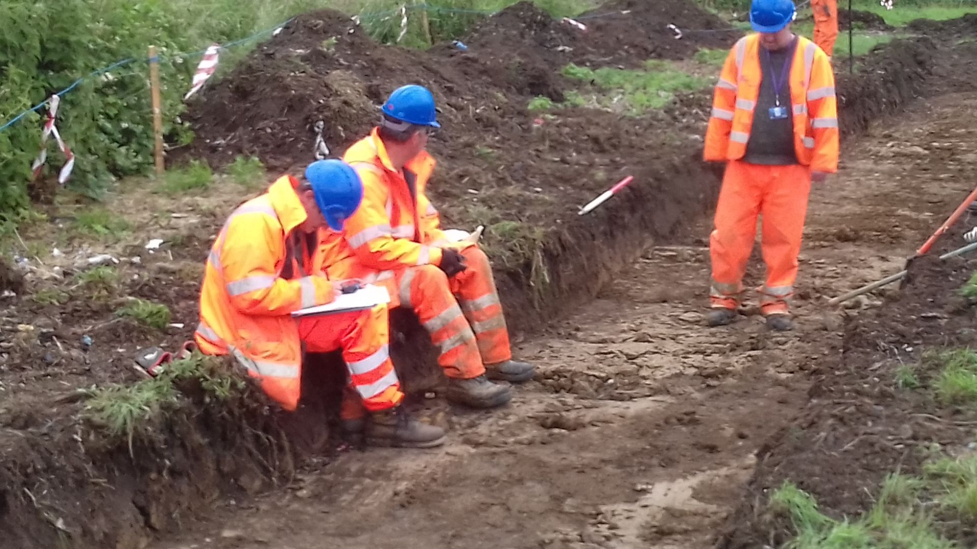 Four archaeologists, two siting on the edge of a trench working on paperwork.