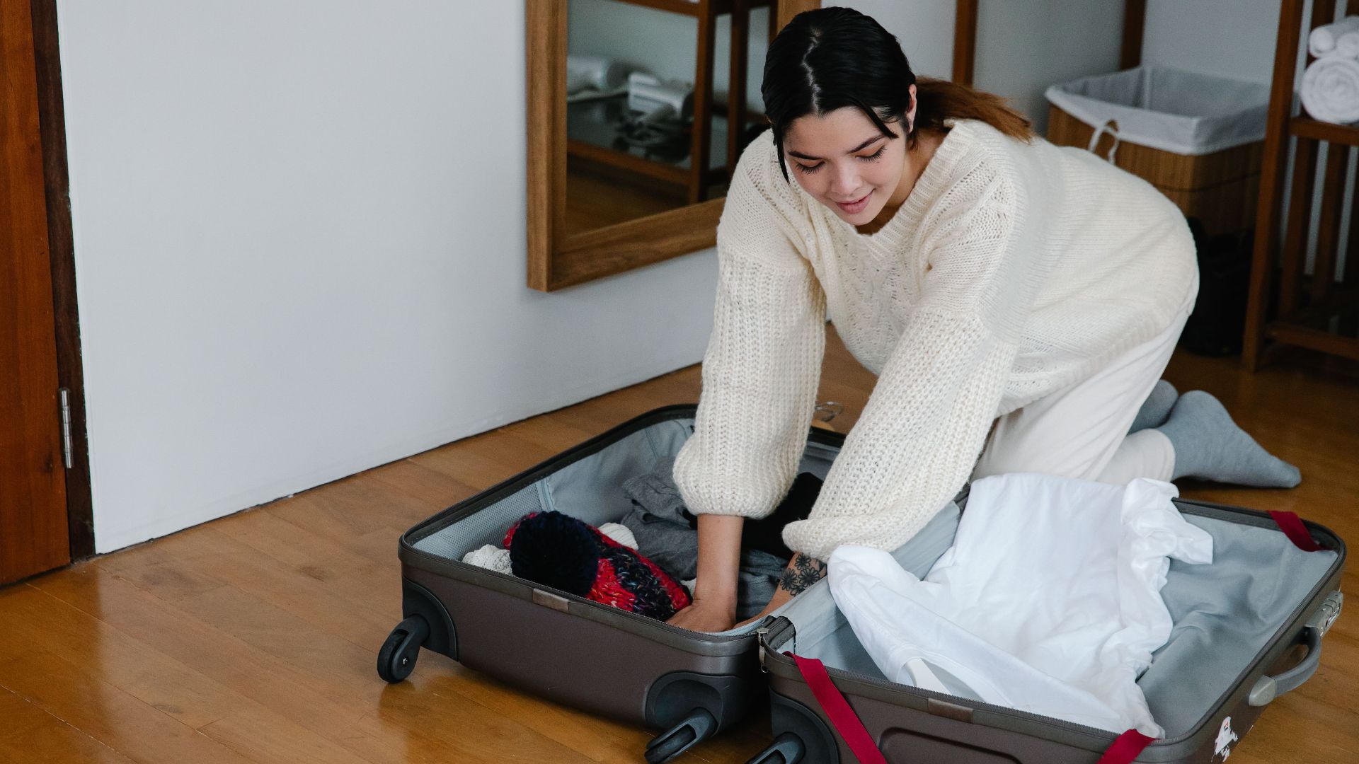 A woman kneels on the floor packing clothes into a suitcase in a cozy bedroom setting.