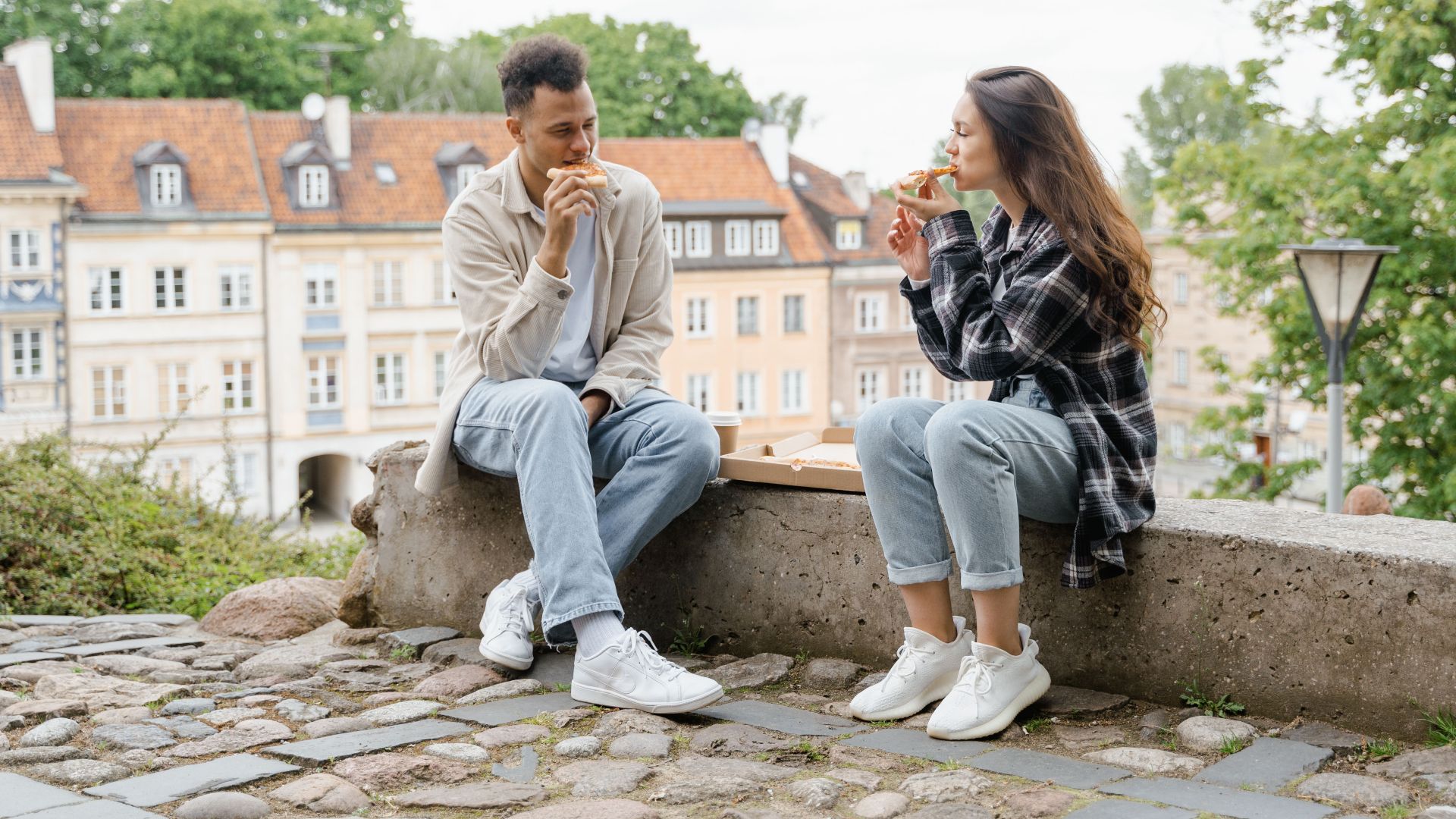 A couple sitting on a bench outdoors, enjoying pizza with picturesque houses in the background.