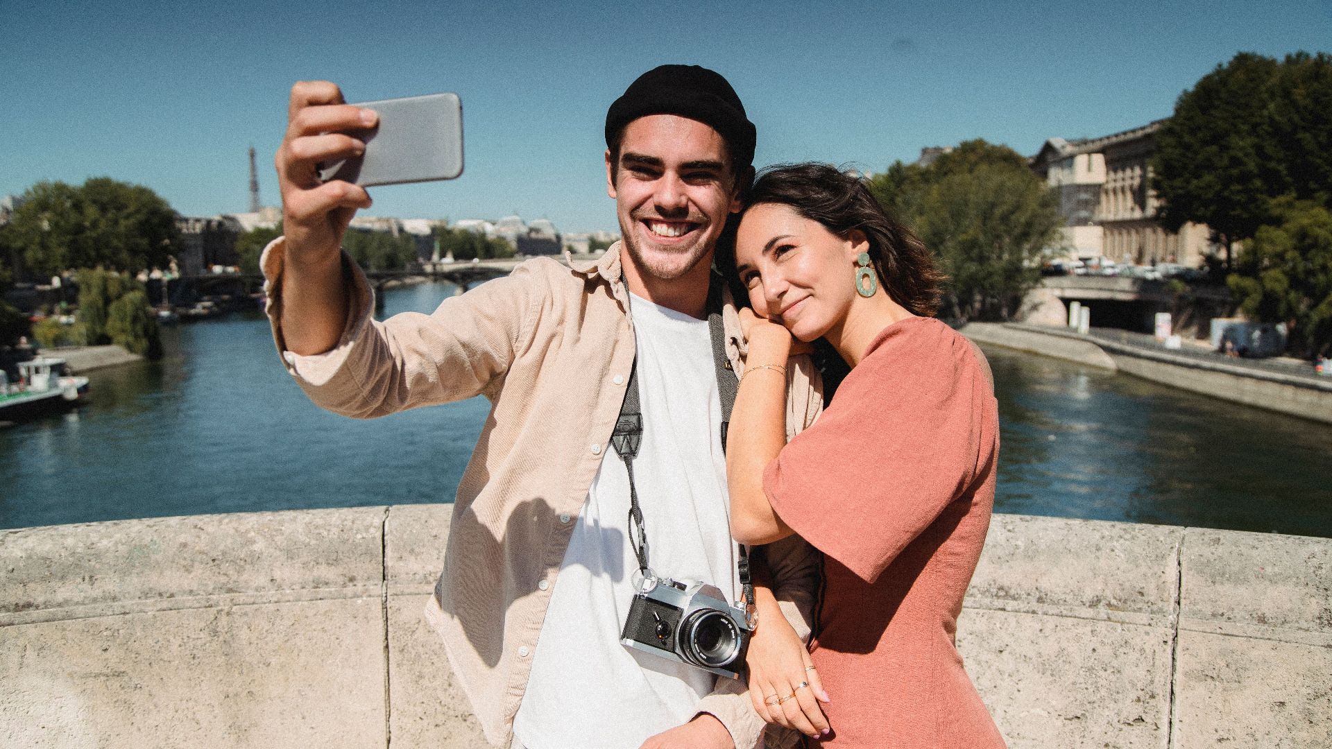 Young couple captures a romantic selfie on a bridge in Paris with scenic background.