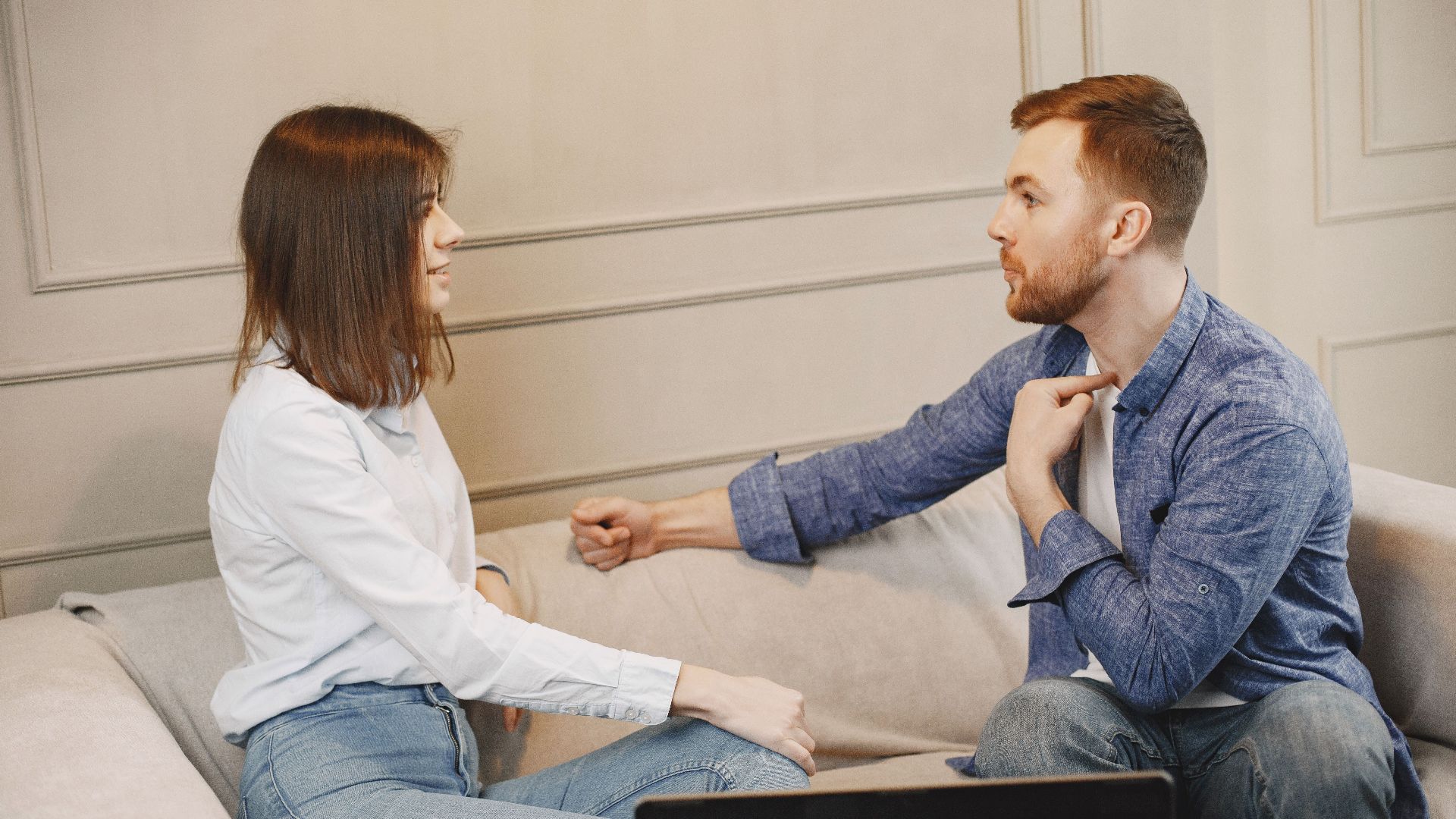 A young couple engaged in a conversation while sitting on a beige couch indoors.