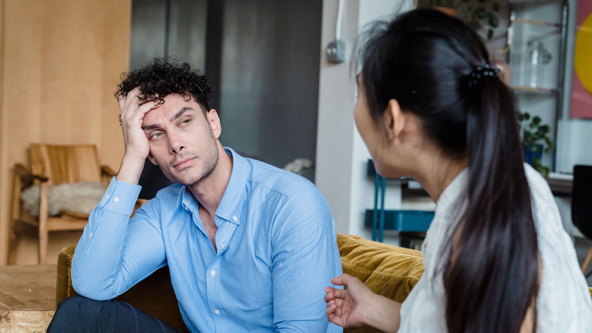 A tired man in a blue shirt listens while a woman talks indoors. Modern living room.