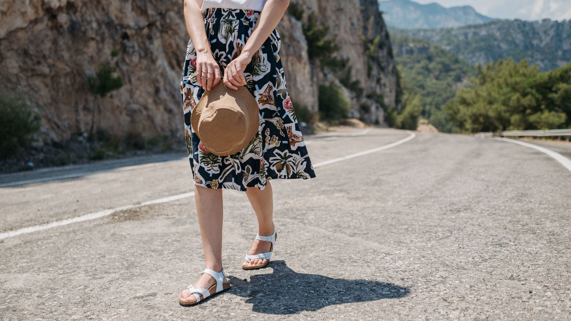 Woman walking along a mountain road wearing a floral skirt and holding a hat, embracing summer vibes.