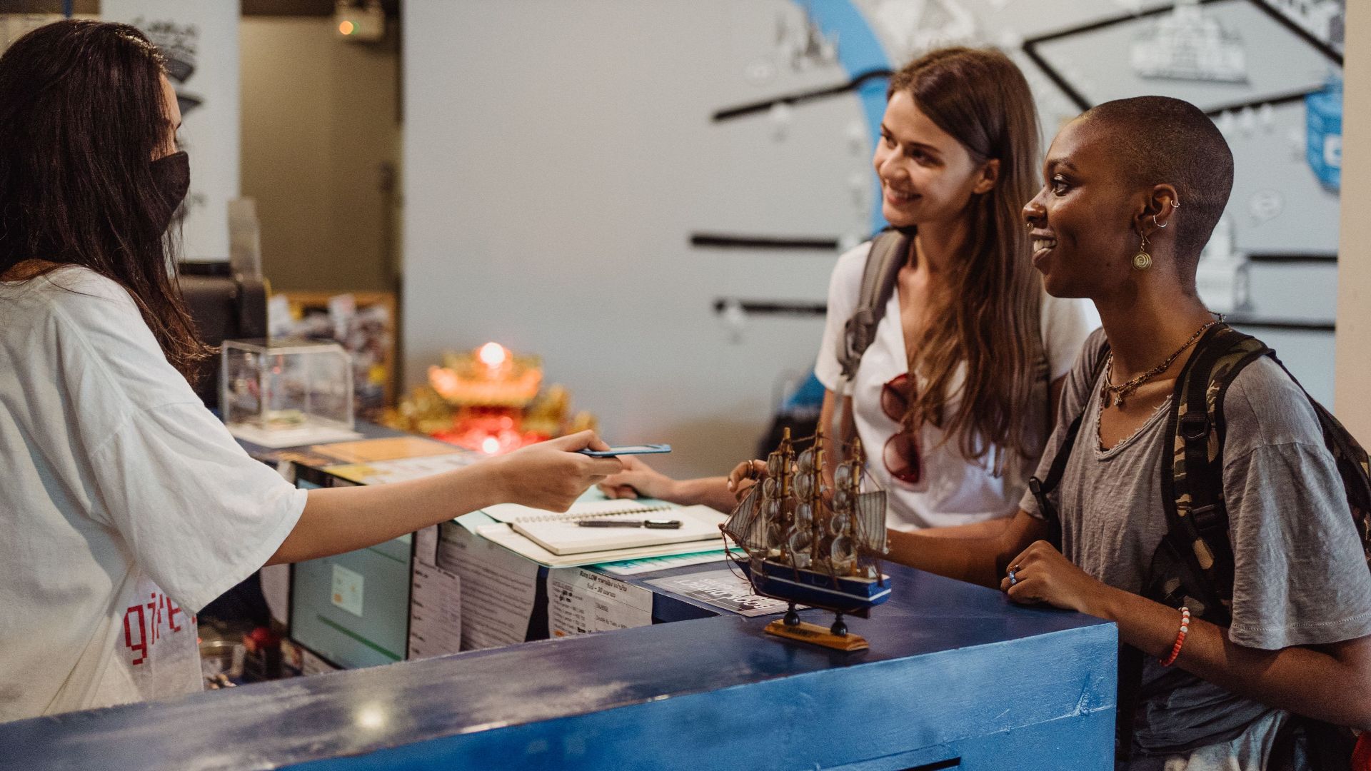 Two women travelers checking in at a hostel reception with a welcoming staff member wearing a face mask.