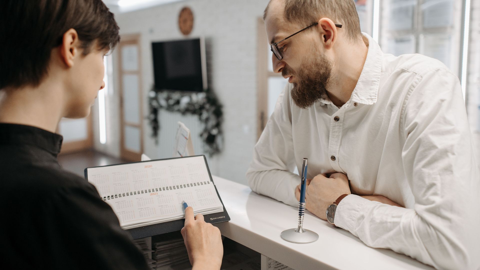 A receptionist and client converse over an appointment book at a clinic reception desk.