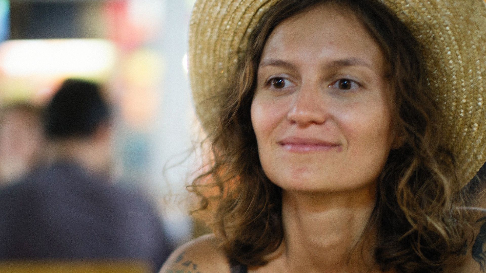 A tattooed woman in a straw hat smiles while sitting outdoors in a relaxed summer setting.