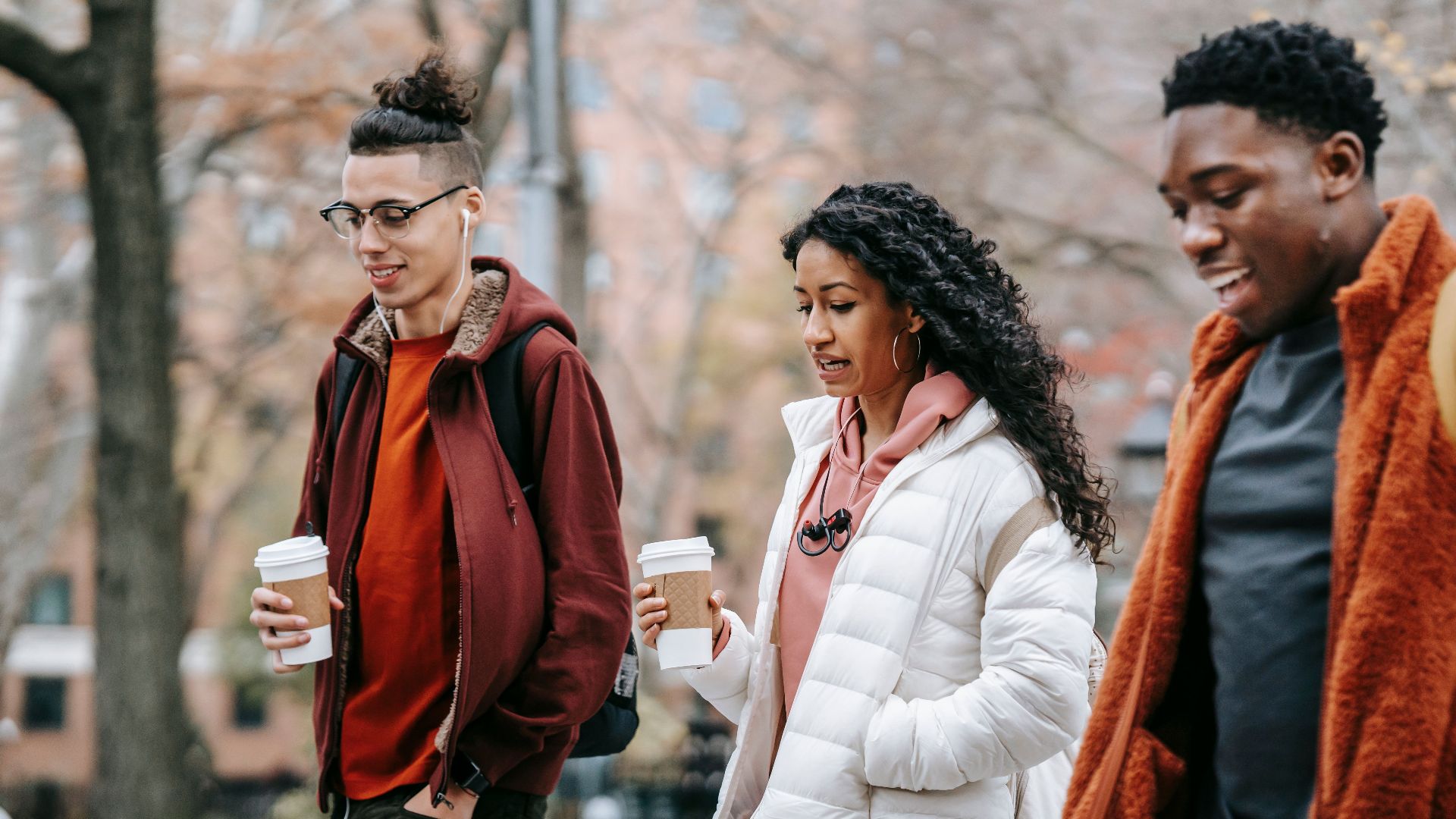 Confident young multiracial friends in outerwear and backpacks talking while strolling in city street near trees with paper cups with drink and earphones in daytime in park