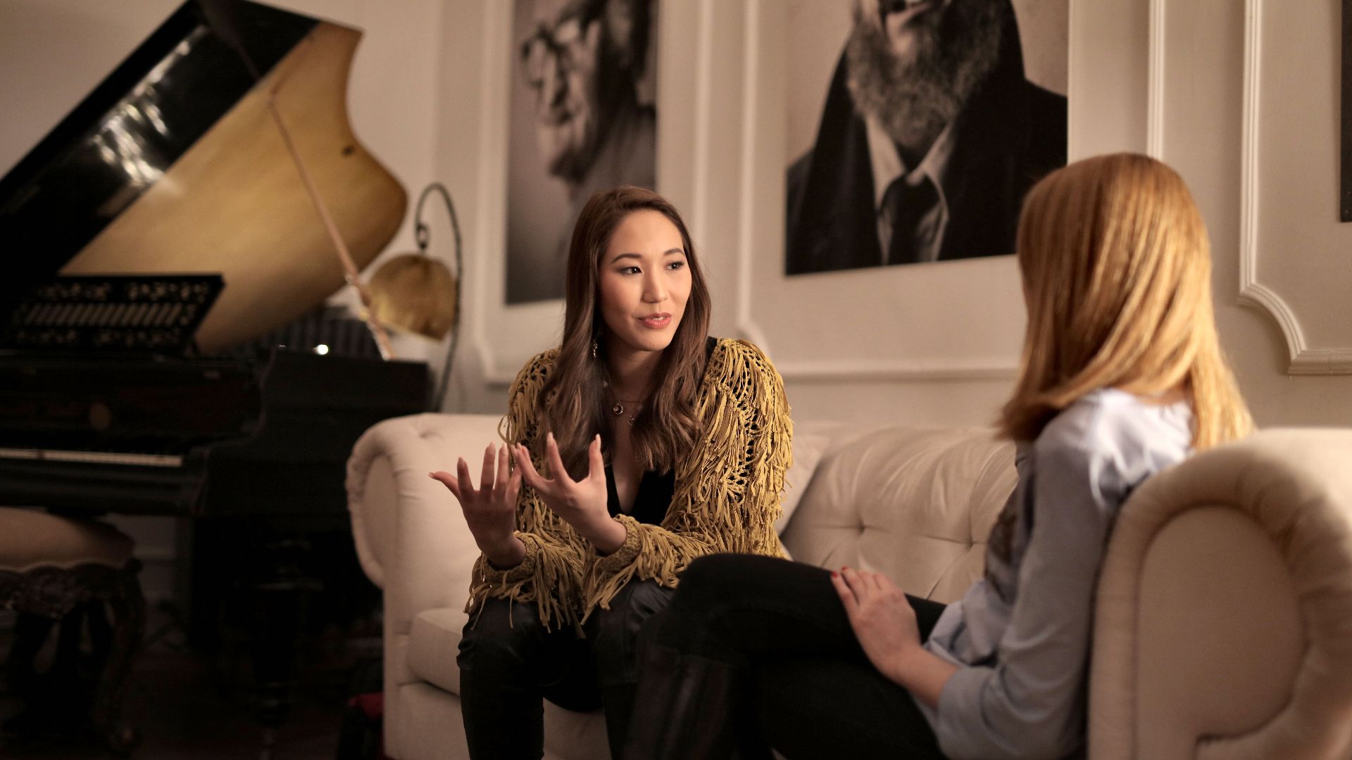 Two women engaging in a relaxed conversation on a couch next to a grand piano in a cozy room.