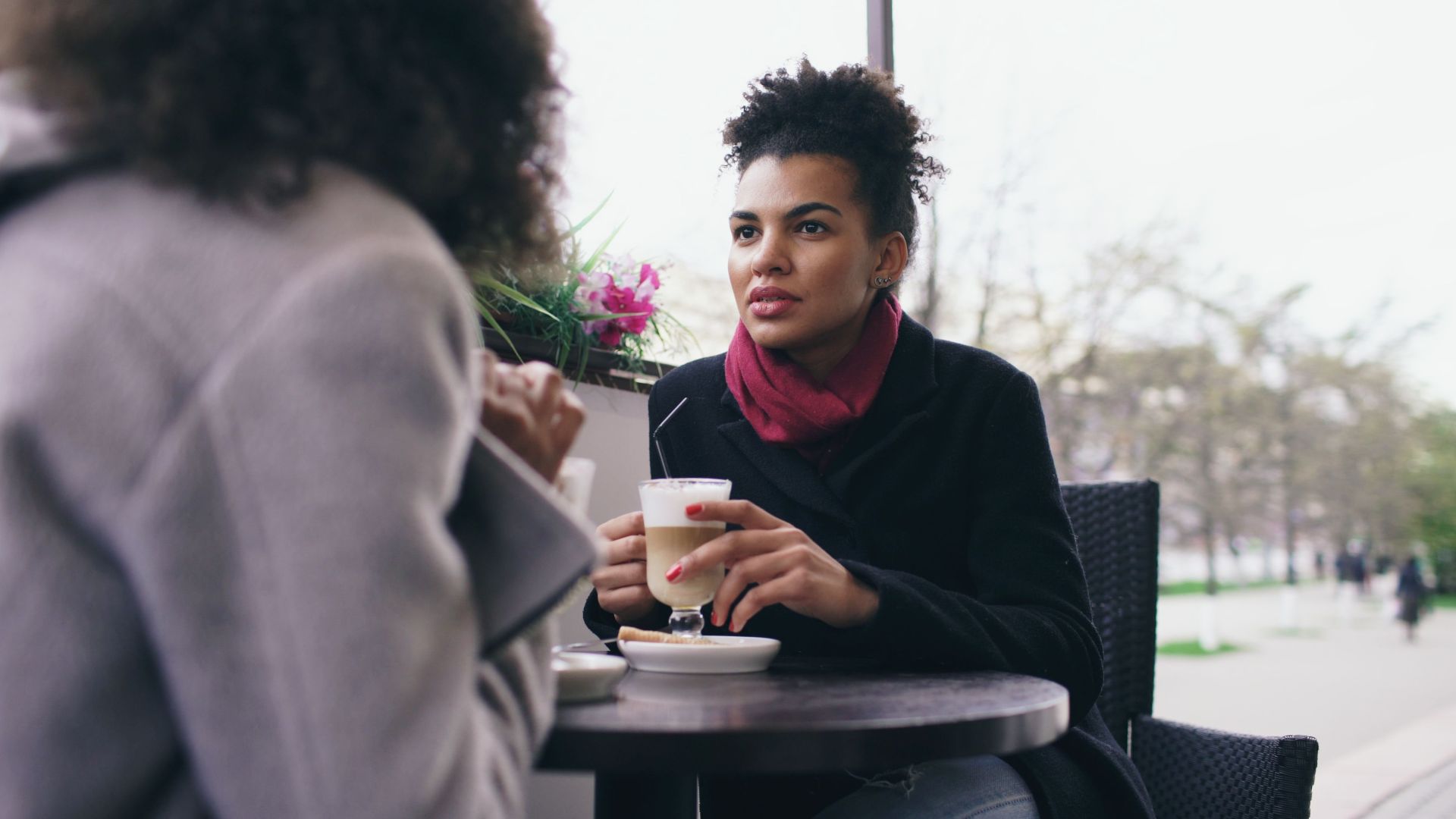 Two women having a conversation over coffee at an outdoor cafe on a bright day.