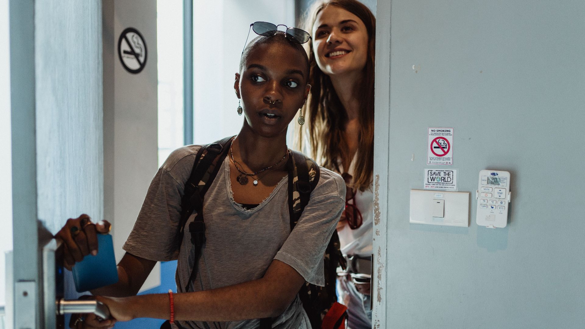 Two young women with backpacks enter a hostel room, ready for adventure.