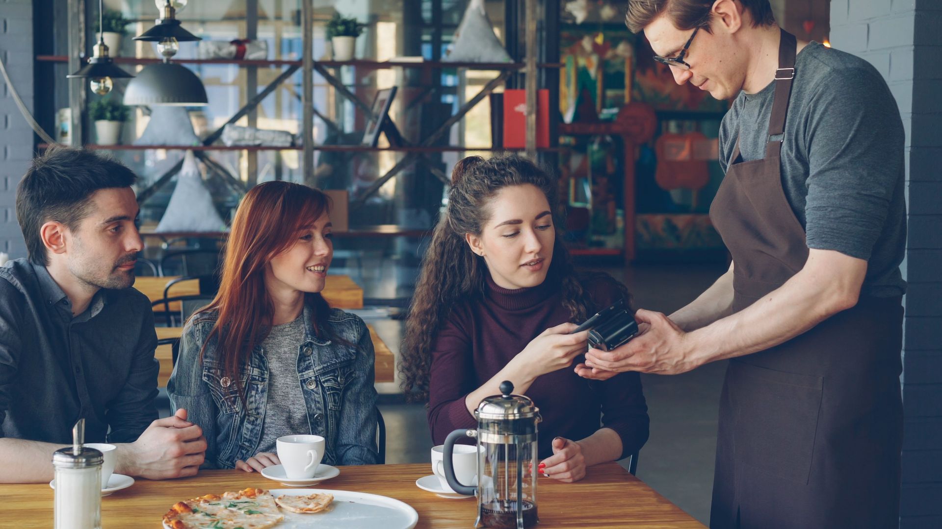 Group of friends having coffee and paying with card in a cozy café setting.