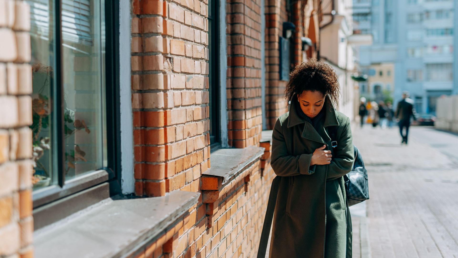 A woman in a green coat walking along a brick building on a sunny day, reflecting solitude.