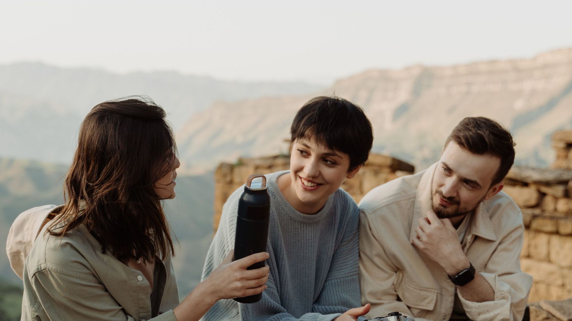 Three friends sit together on a stone wall, enjoying a scenic mountain view in the Caucasus region.