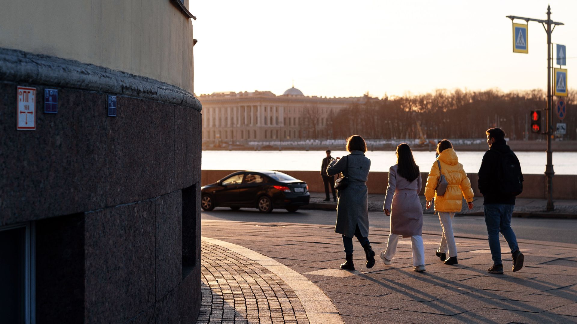 People crossing a city street near a river at sunset, showcasing urban life and architecture.