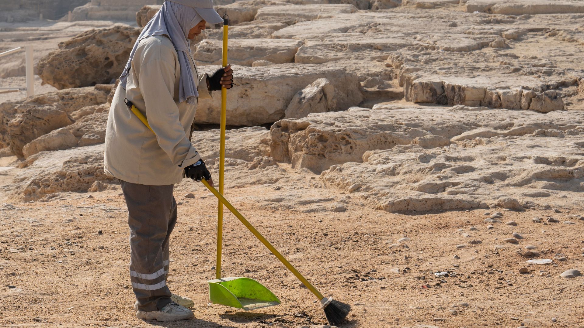 A person cleaning sand near rocky formations during the day, showcasing a desert landscape.