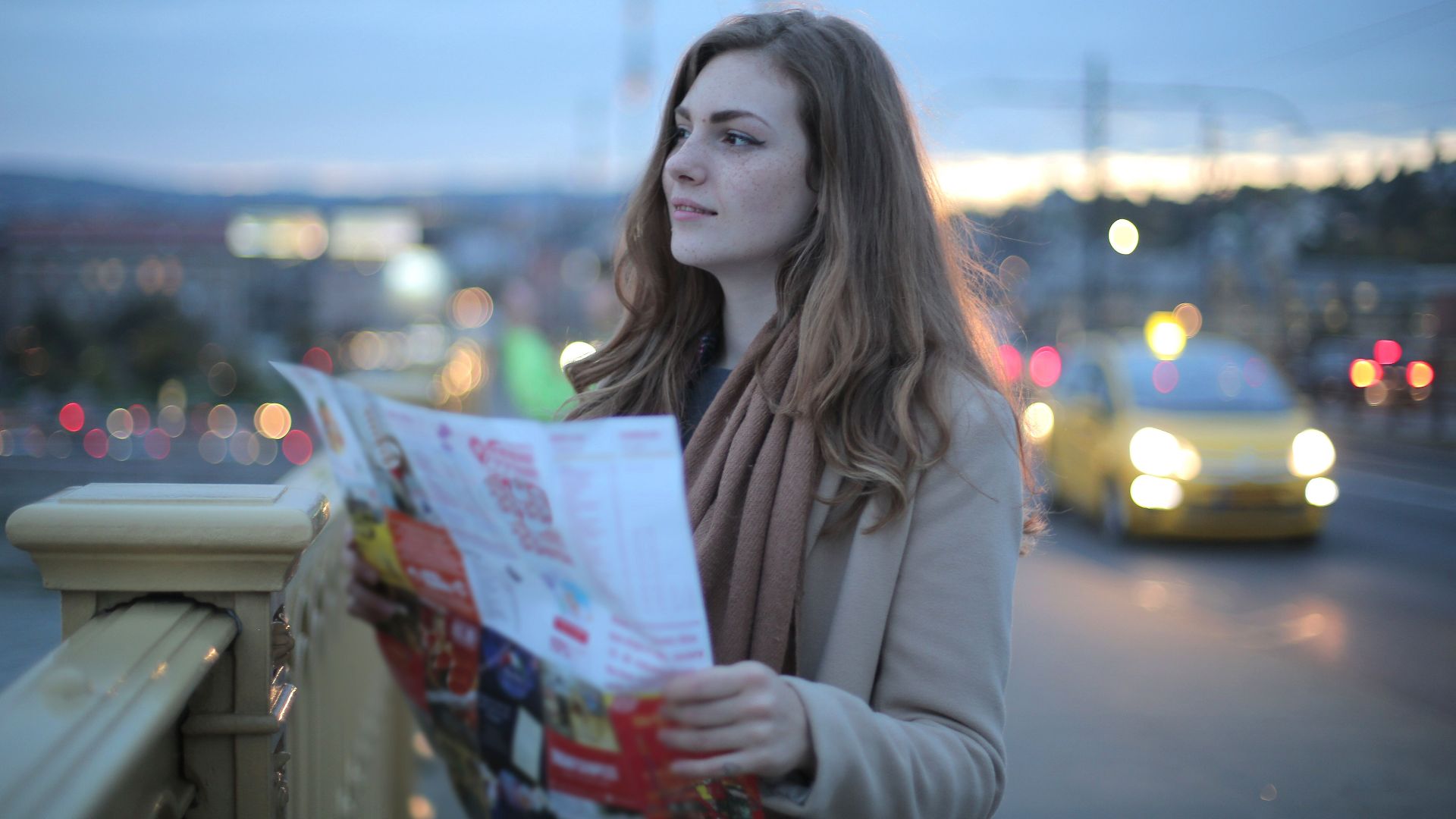 Female traveler in warm clothes standing on street with map and looking away at dusk
