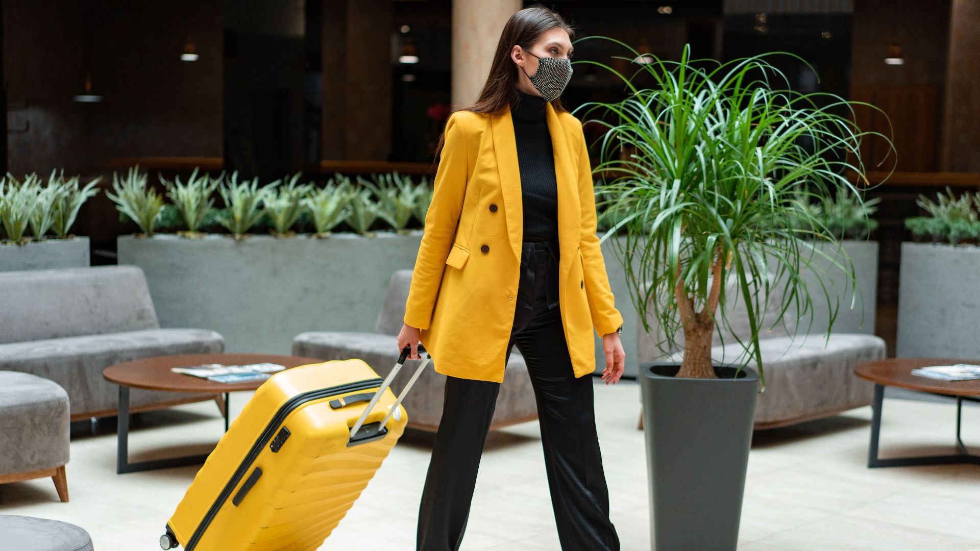 Fashionable woman in yellow blazer and face mask walking with suitcase indoors.