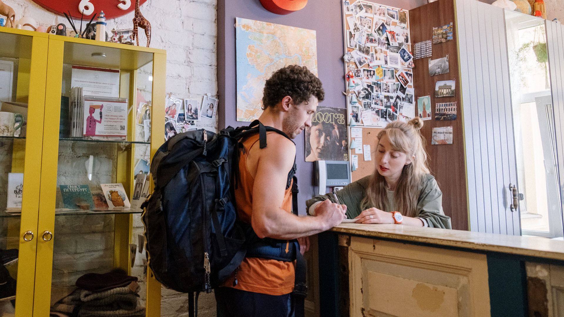 A backpacker at a hostel front desk getting assistance from a receptionist.